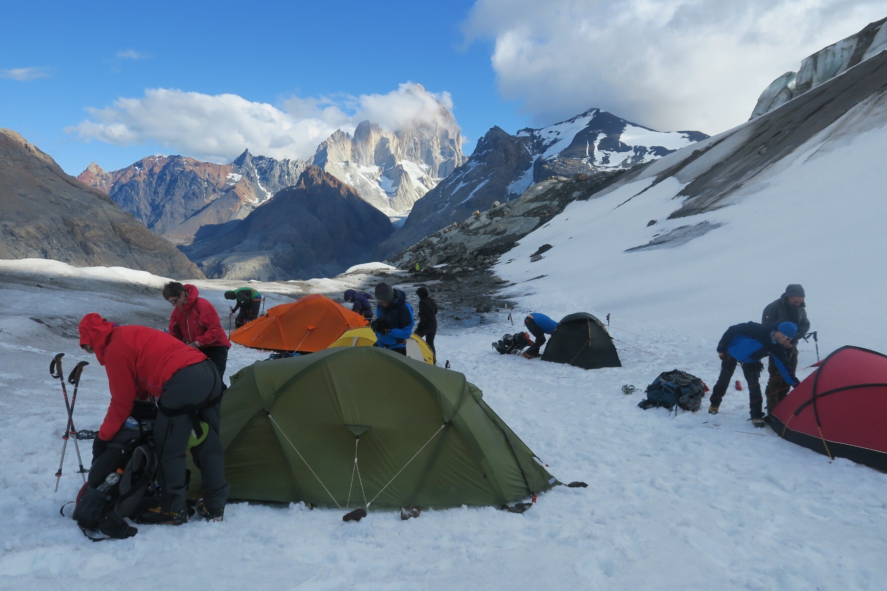 Some tents are being prepared in the snow in the foreground. The background has some jagged mountain peaks with clouds