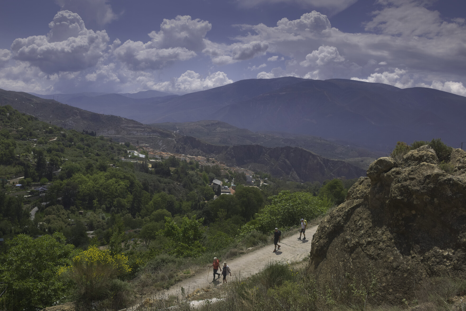 Some hikers on a dirt track in the foreground. In the middle distance lies a town. Behind are a range of mountains with clouds above them