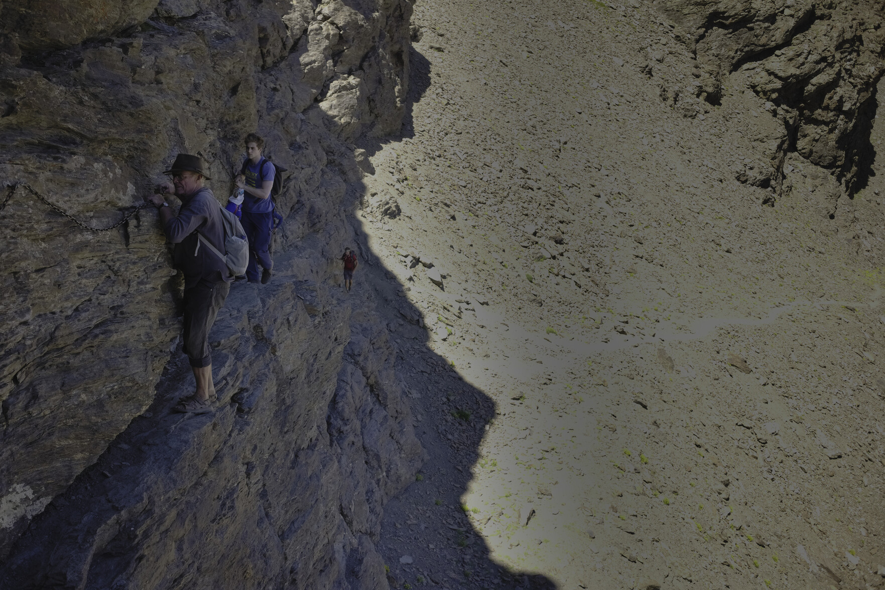 A man on the left moves slowly across a very narrow rocky ledge whilst holding onto a chain fixed to the rock wall. To the right the rock falls away vertically to the valley floor