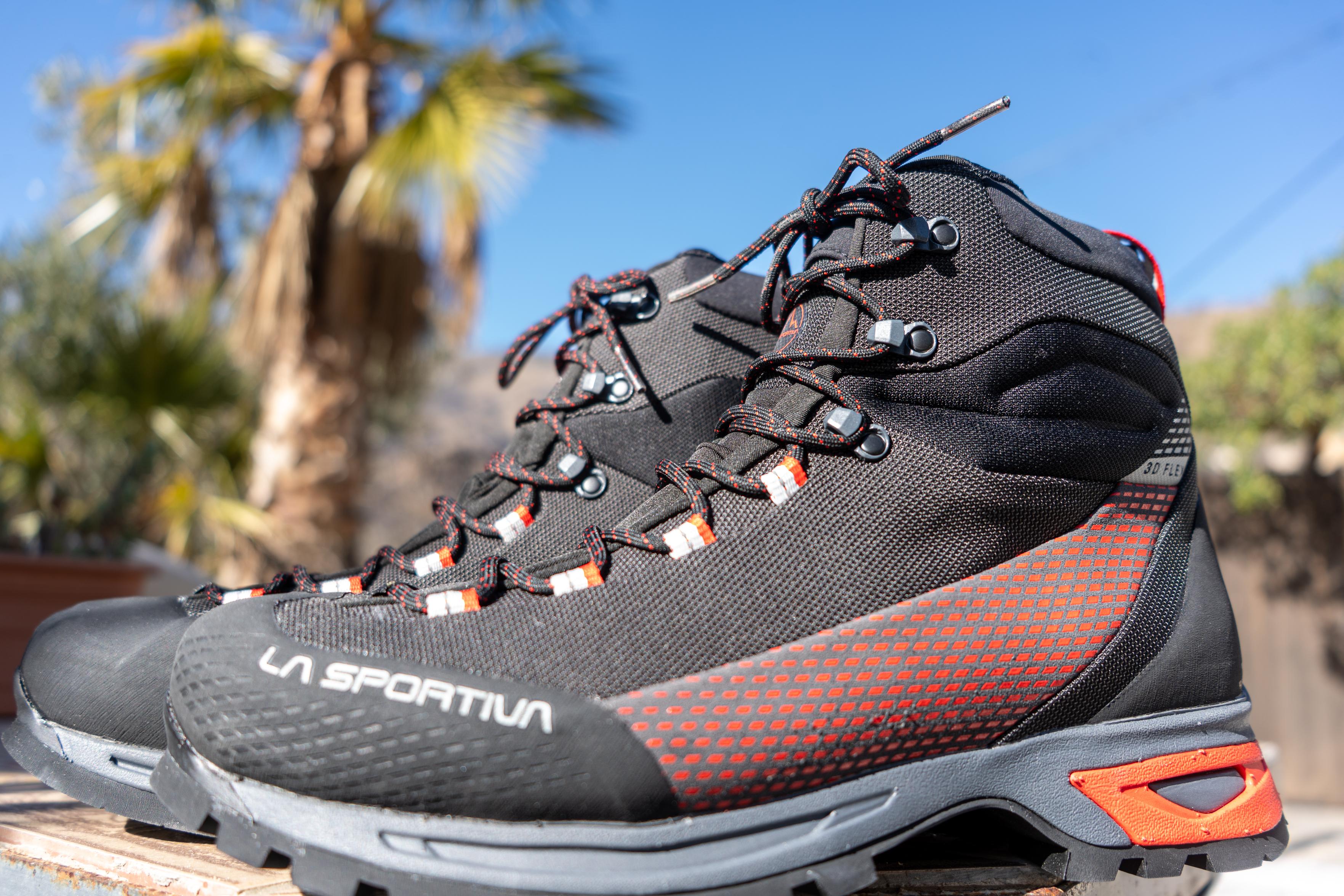 A pair of hiking boots from La Sportiva stand side by side on a table. Above is blue sky and a palm tree