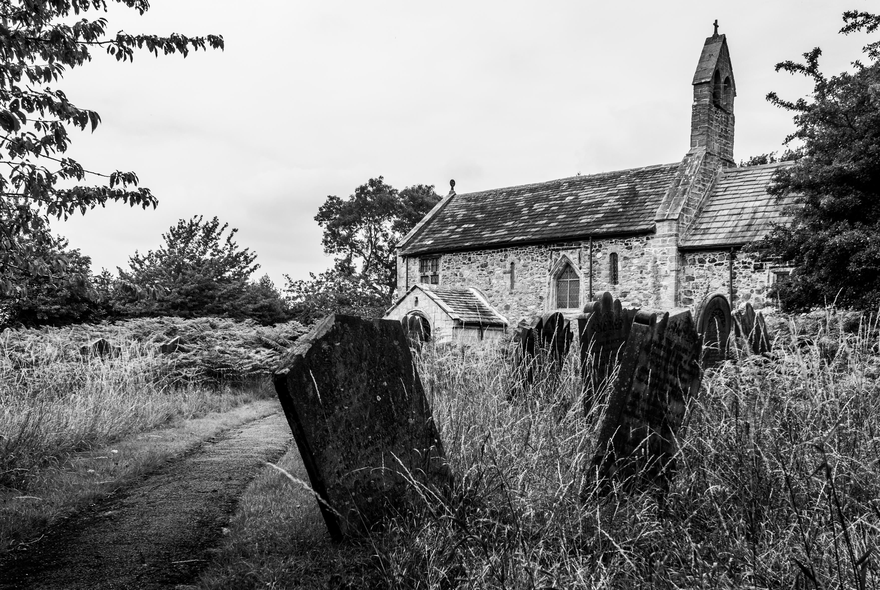 Black and white image of an old church and leaning headstones with tall overgrown vegetation