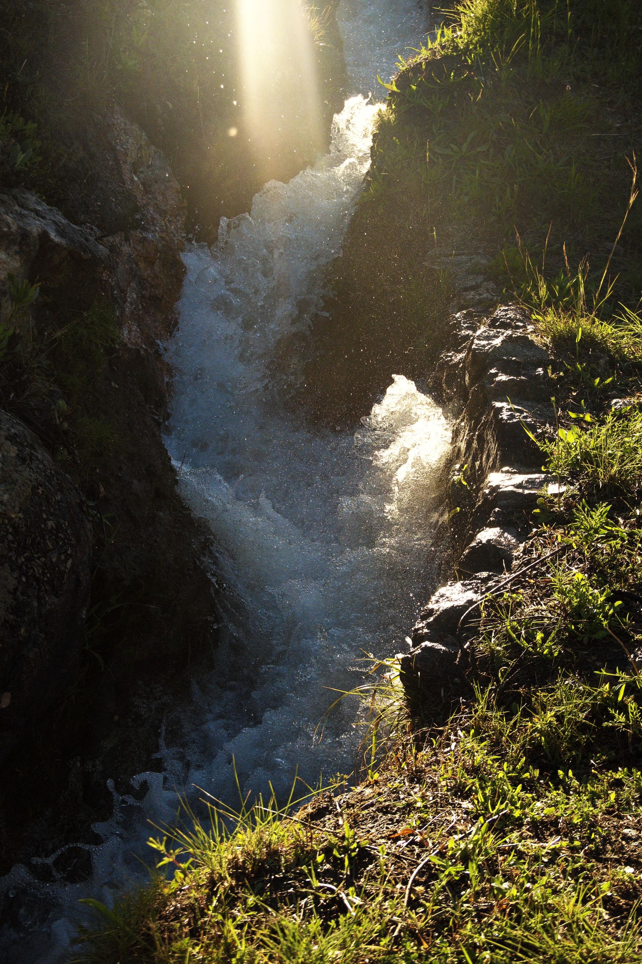 Green grass on the right hand bank alongside a narrow water channel with fast flowing water. Light teems through from the top and illuminates some of the water