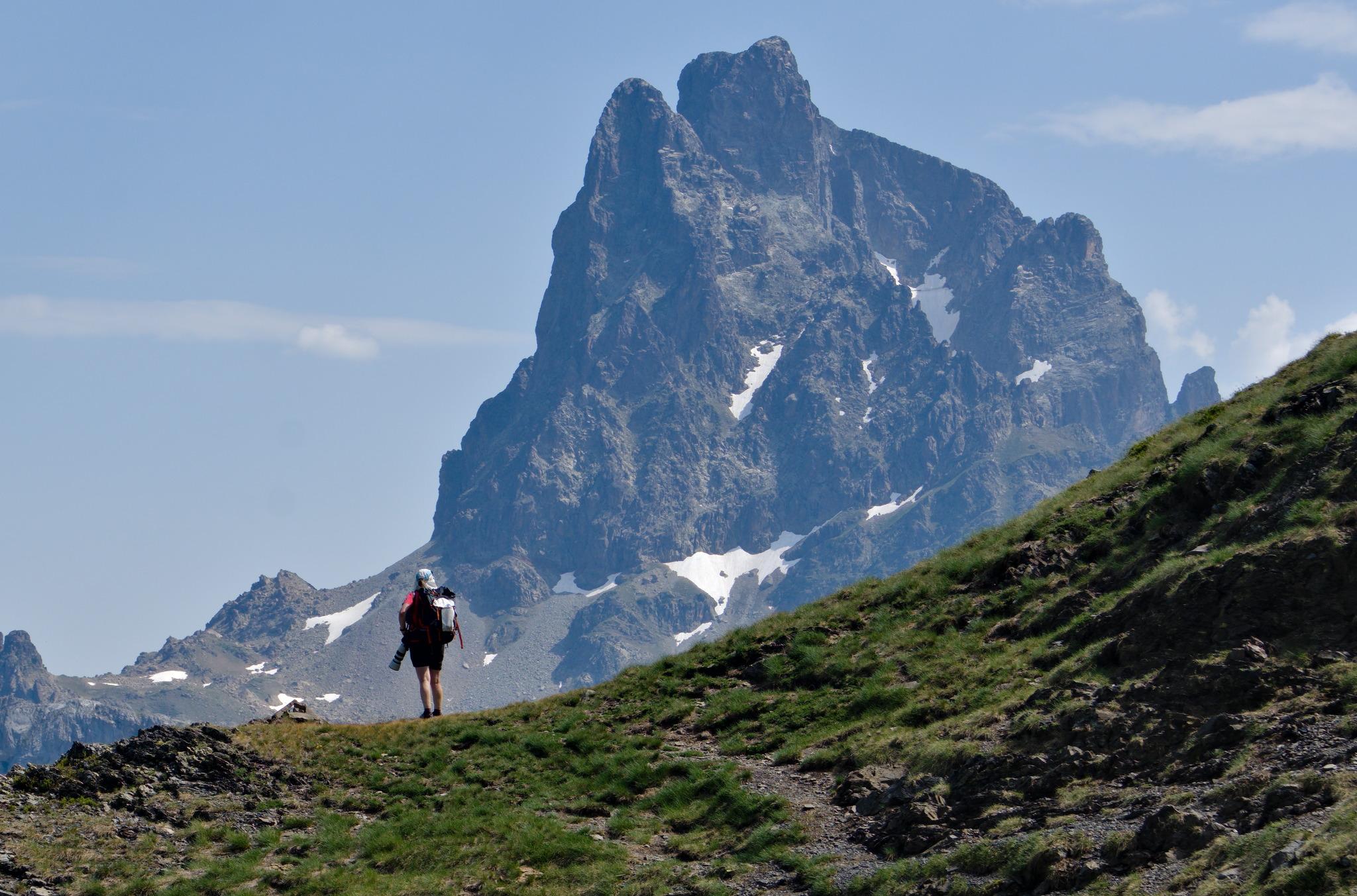 A person crosses a grassy hillside along a trail. Behind rises a huge rocky mountain.