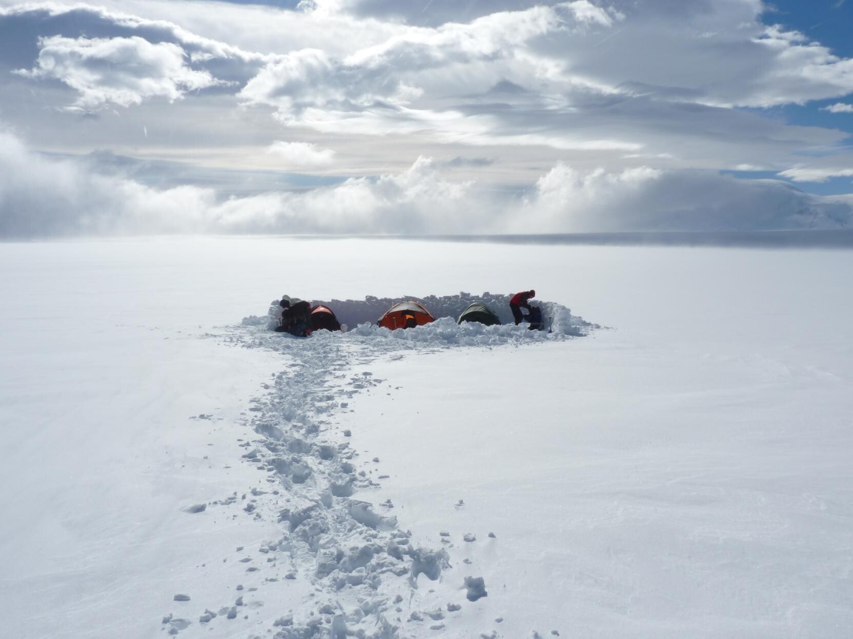A small group of tents sits behind a snow wall in the middle of a white snow filled wilderness