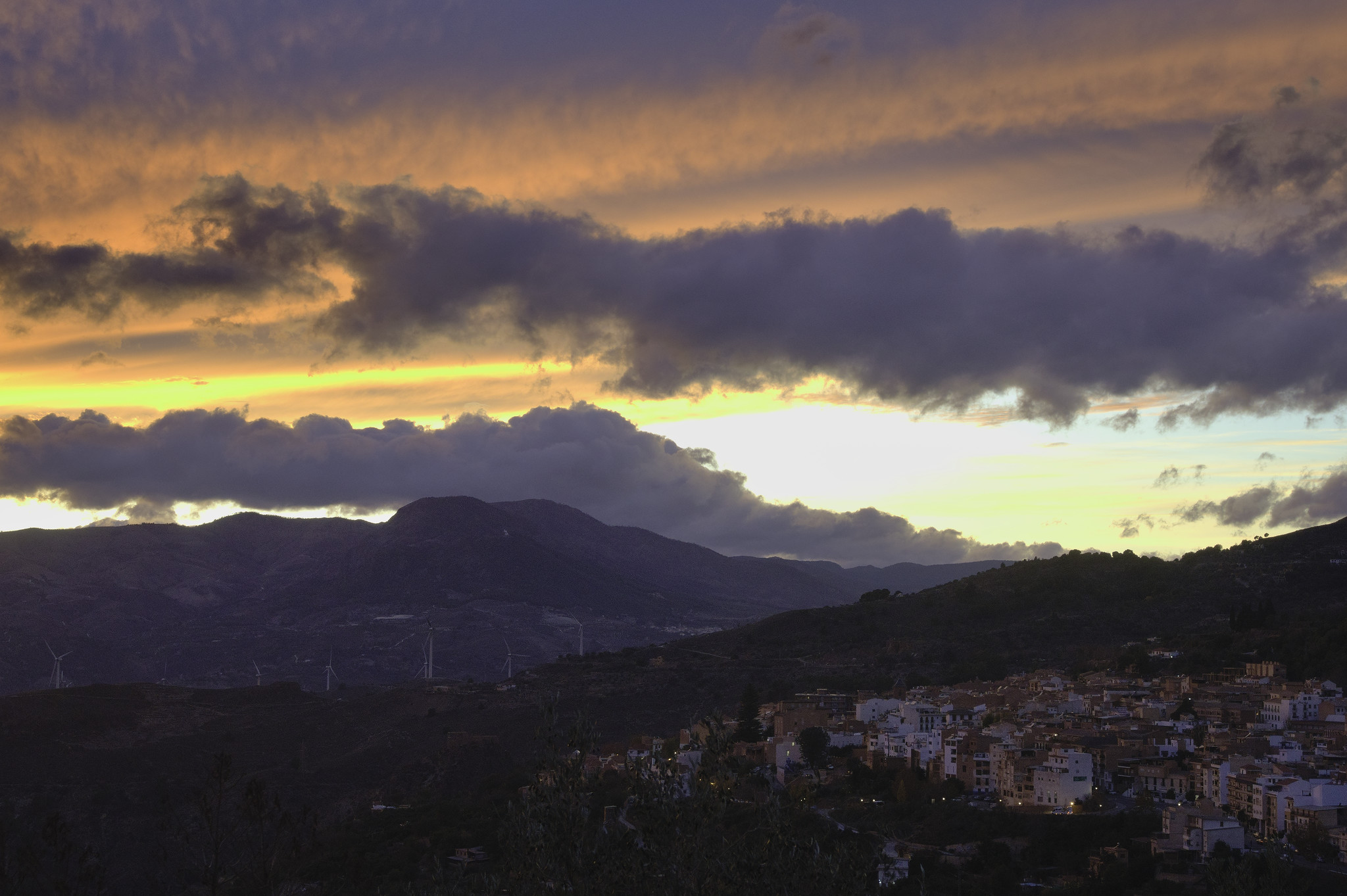 Bottom left is the outline of a spanish town at dusk. Above some mountains and dark clouds surrounded by a colorful sunset of orange and yellow