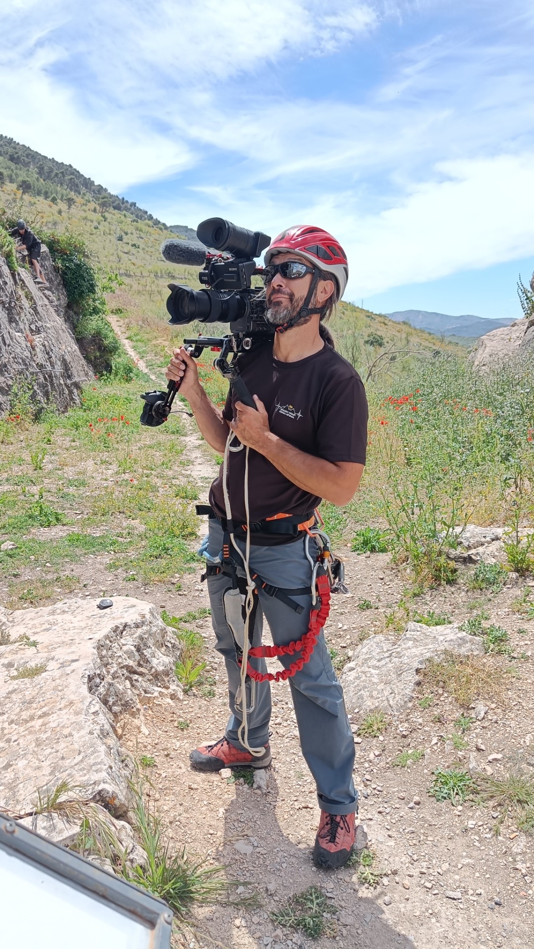 Person with helmet, harness and via ferrata equipment holds camera filming gear