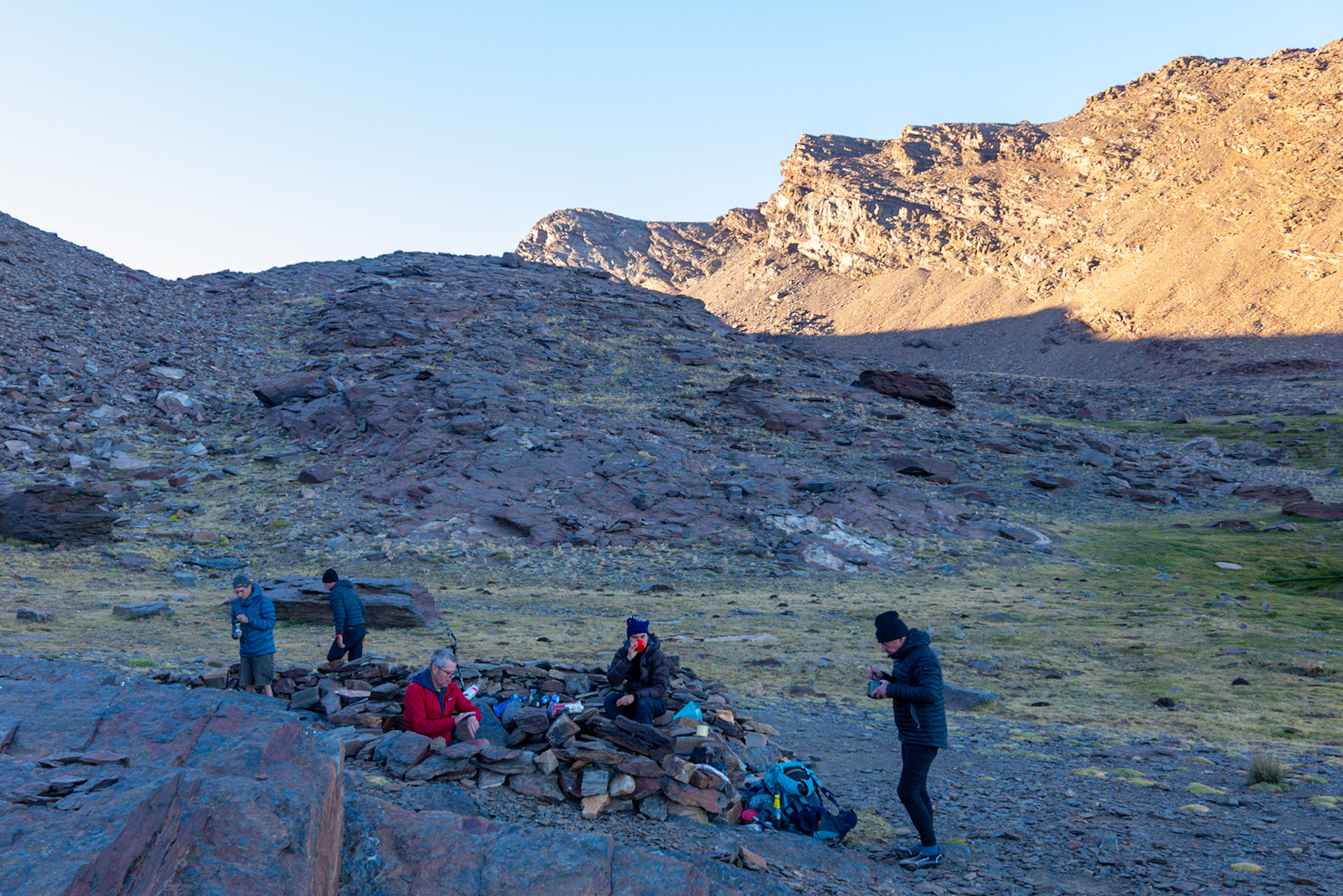 A group of backpackers are preparing an evening meal in small walled shelters. Behind the final rays of the sun reach the mountain of Alcazaba 3269m