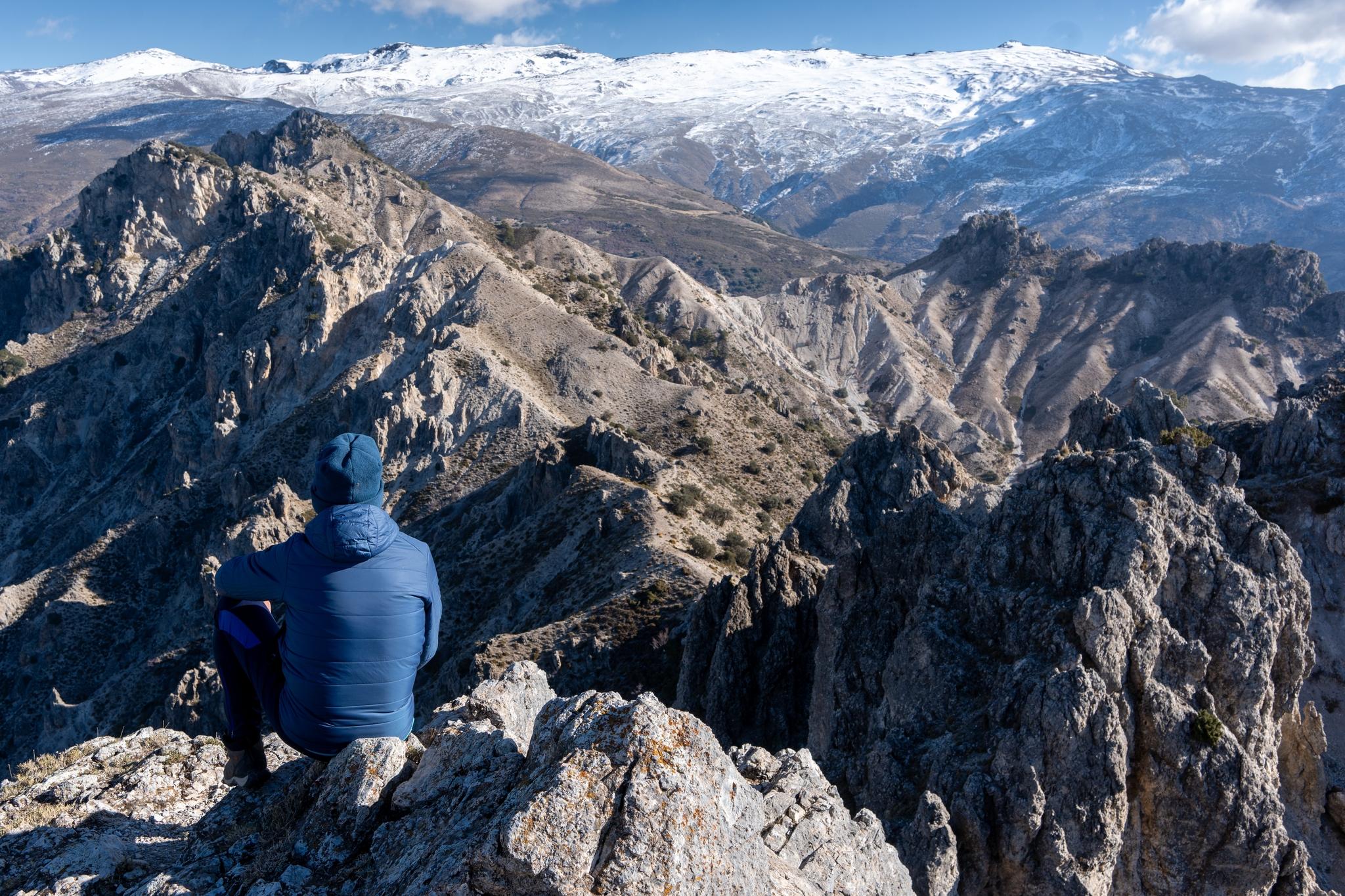 A person is sat at the summit of a mountain with a jagged series of peaks stretching into the distance. Beyond rises some even higher snow clad mountains