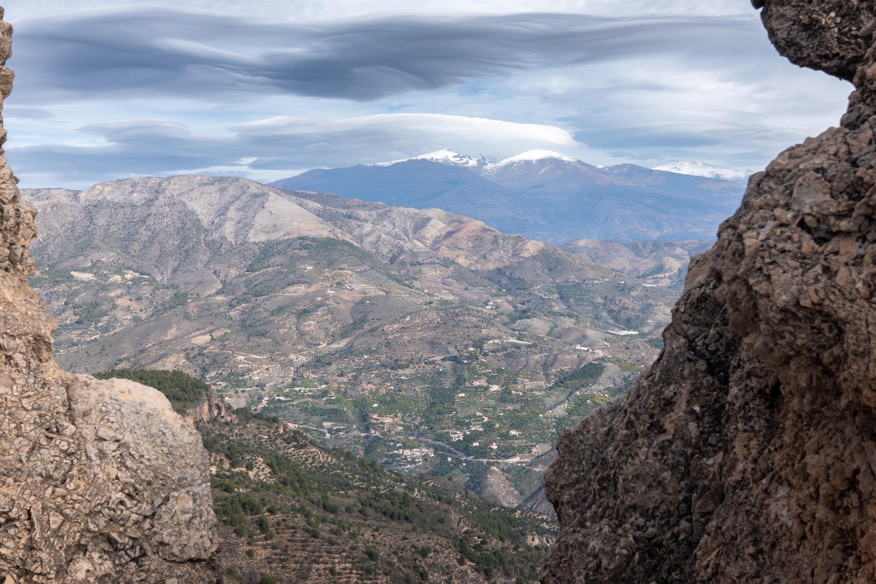 A narrow gap in the rock wall gives a view past green hills and valleys to the snow clad peaks beyond. Some dark threatening clouds above
