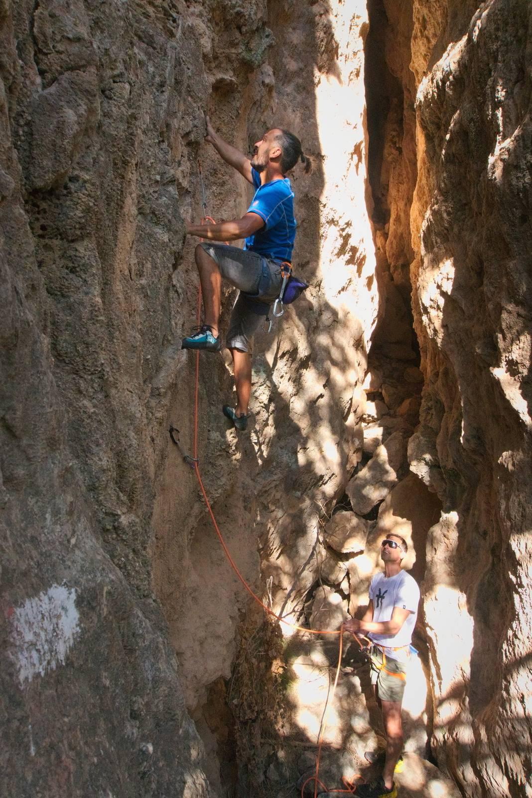 A person in blue climbs a steep rock wall on the left belayed by a person below with white t-shirt (Kenton)