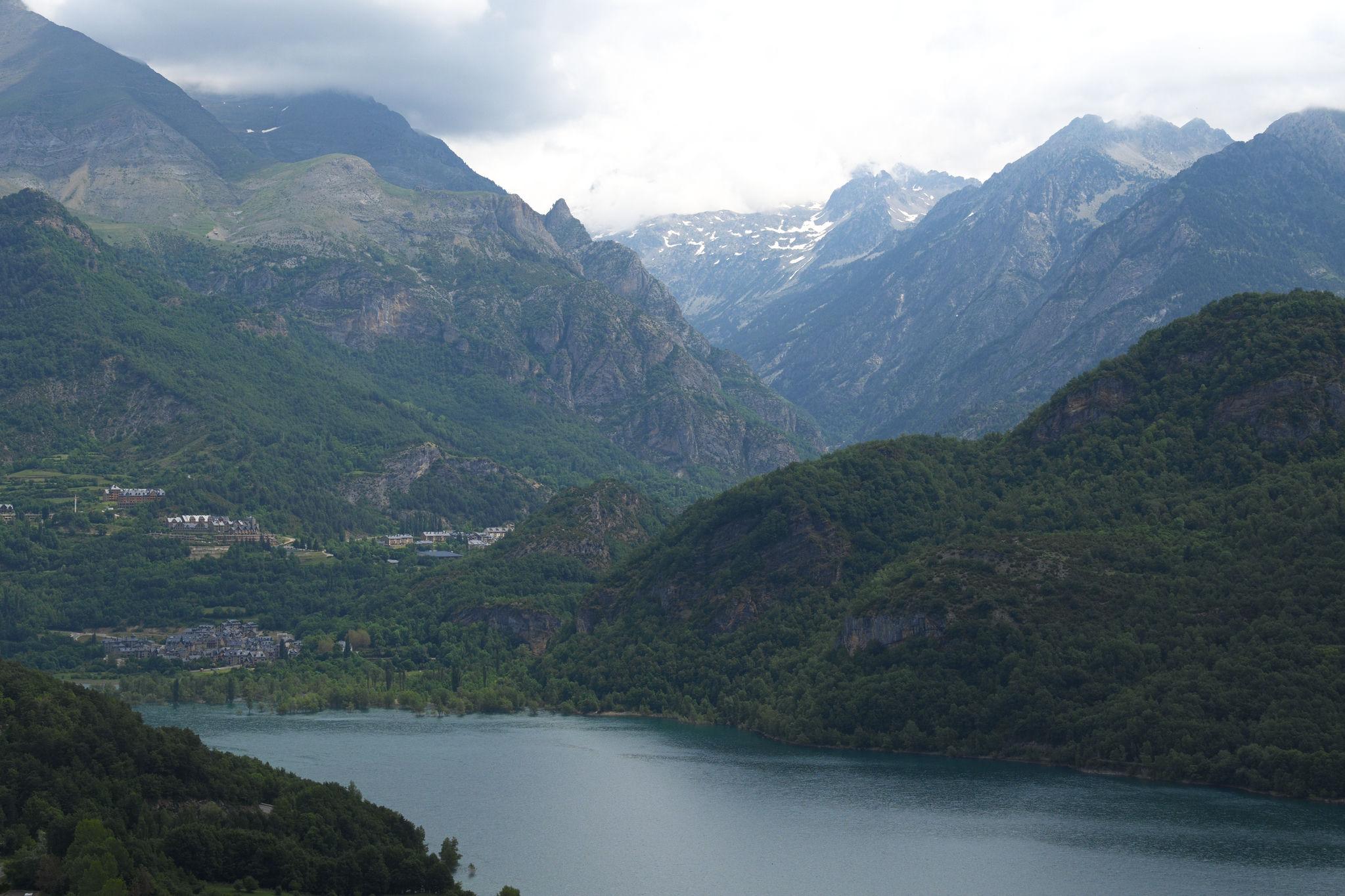 A town stands on the far shore of a lake. Lake is surrounded by lush forests. A deep valley leads to high mountains with snow