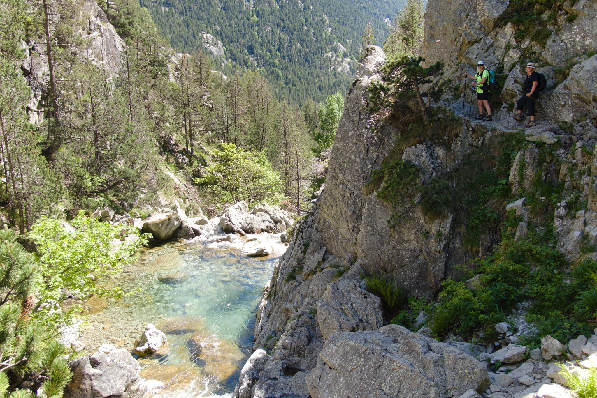A narrow track on the right high above a river valley. The river has deep rock pools. Two people stand on the trail