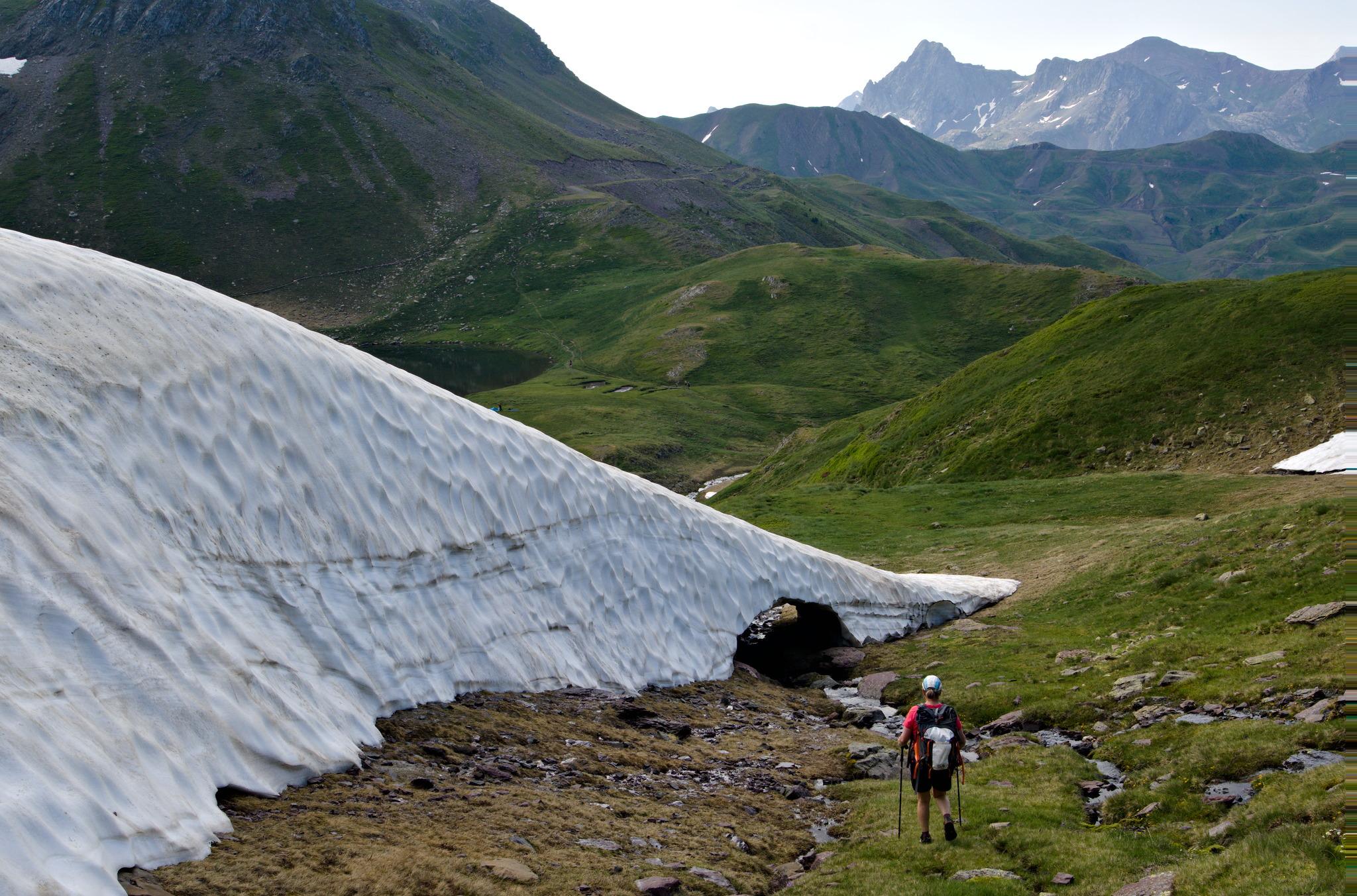 A huge snow bank leads down to a small hole through the snow through which a stream flows. Green hillsides beyond.