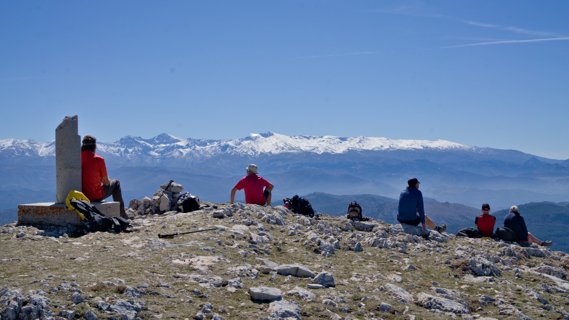 People sitting on a mountain summit with distant views to a snow clad mountain range.