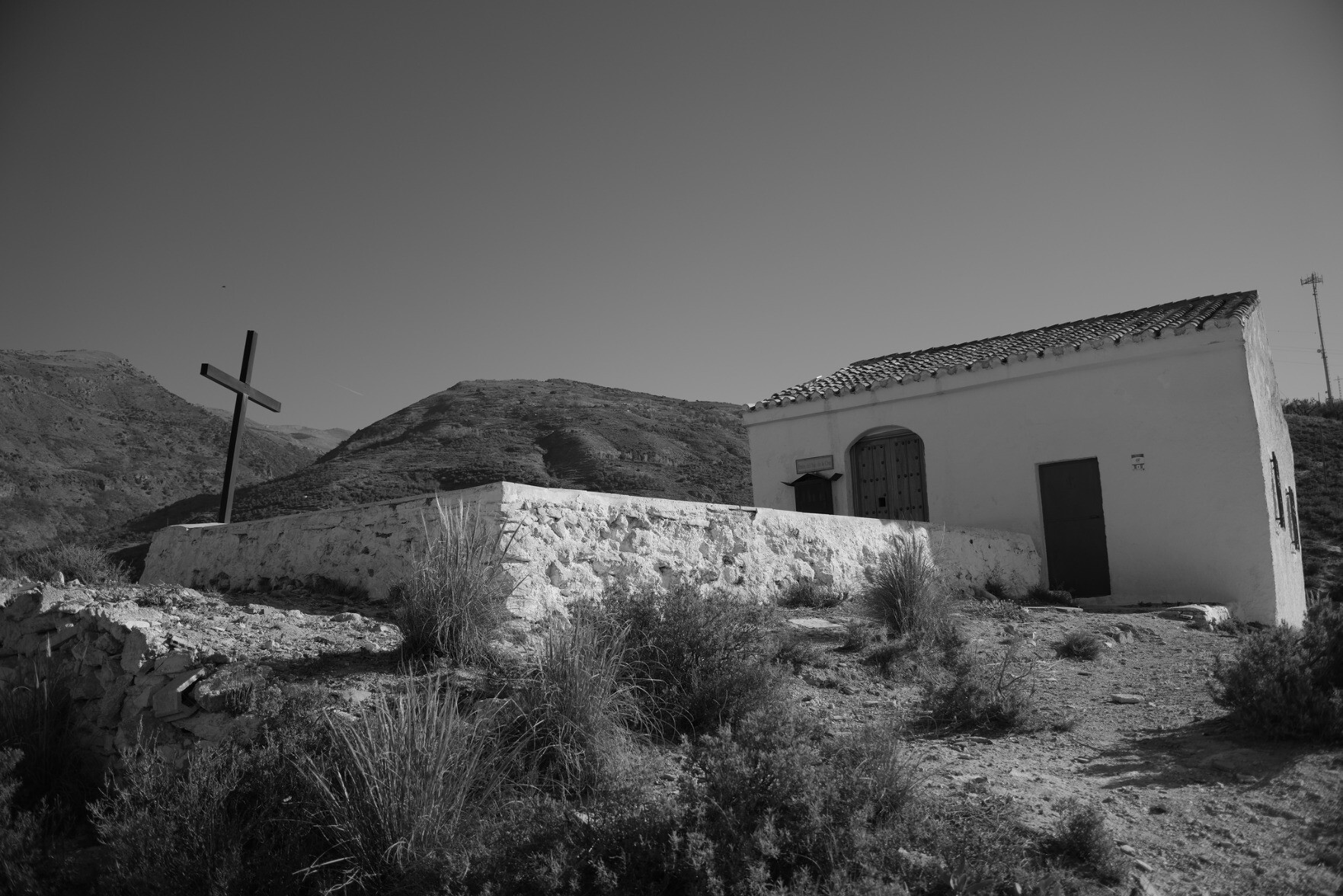 Black and white photo of a small Ermita (chapel) on a hill in Spain. A cross is to the left and a small chapel to the right