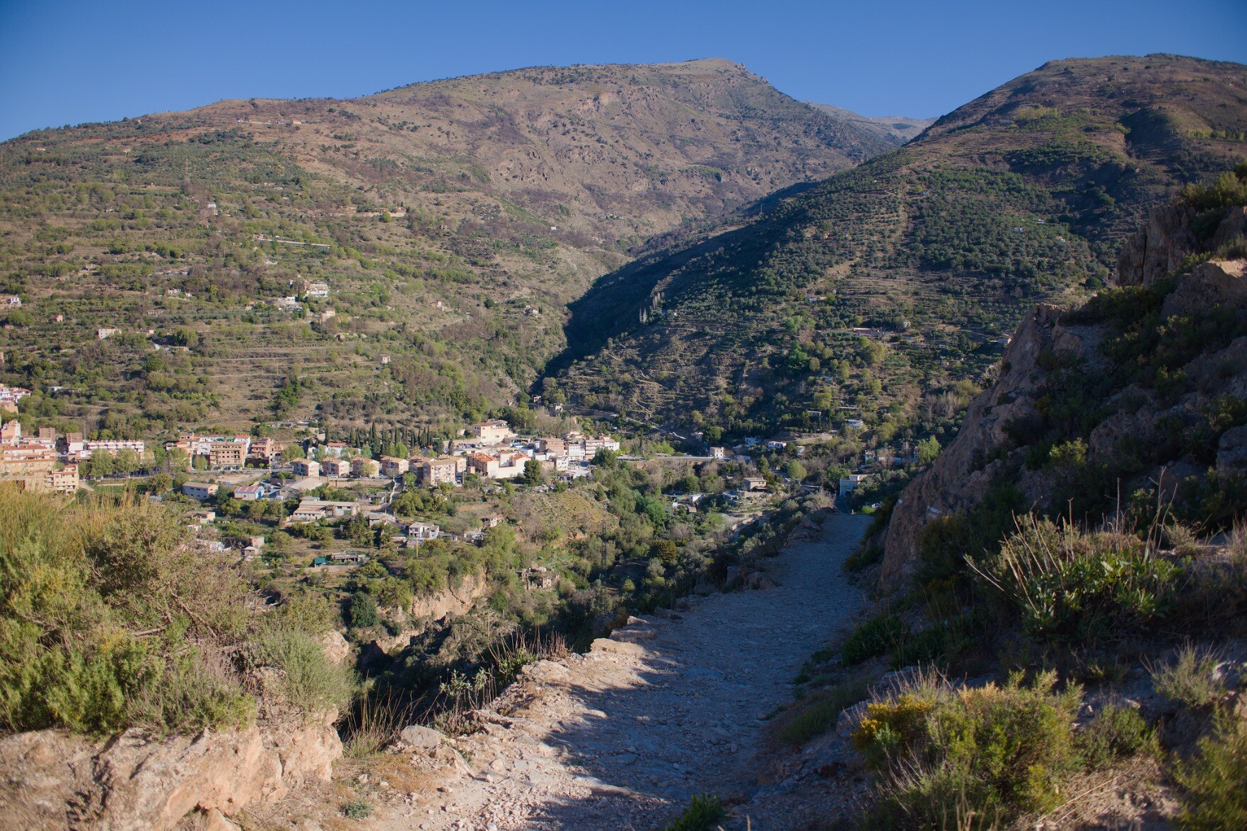 A path leads down to the right whilst behind is a spanish town with mountains behind