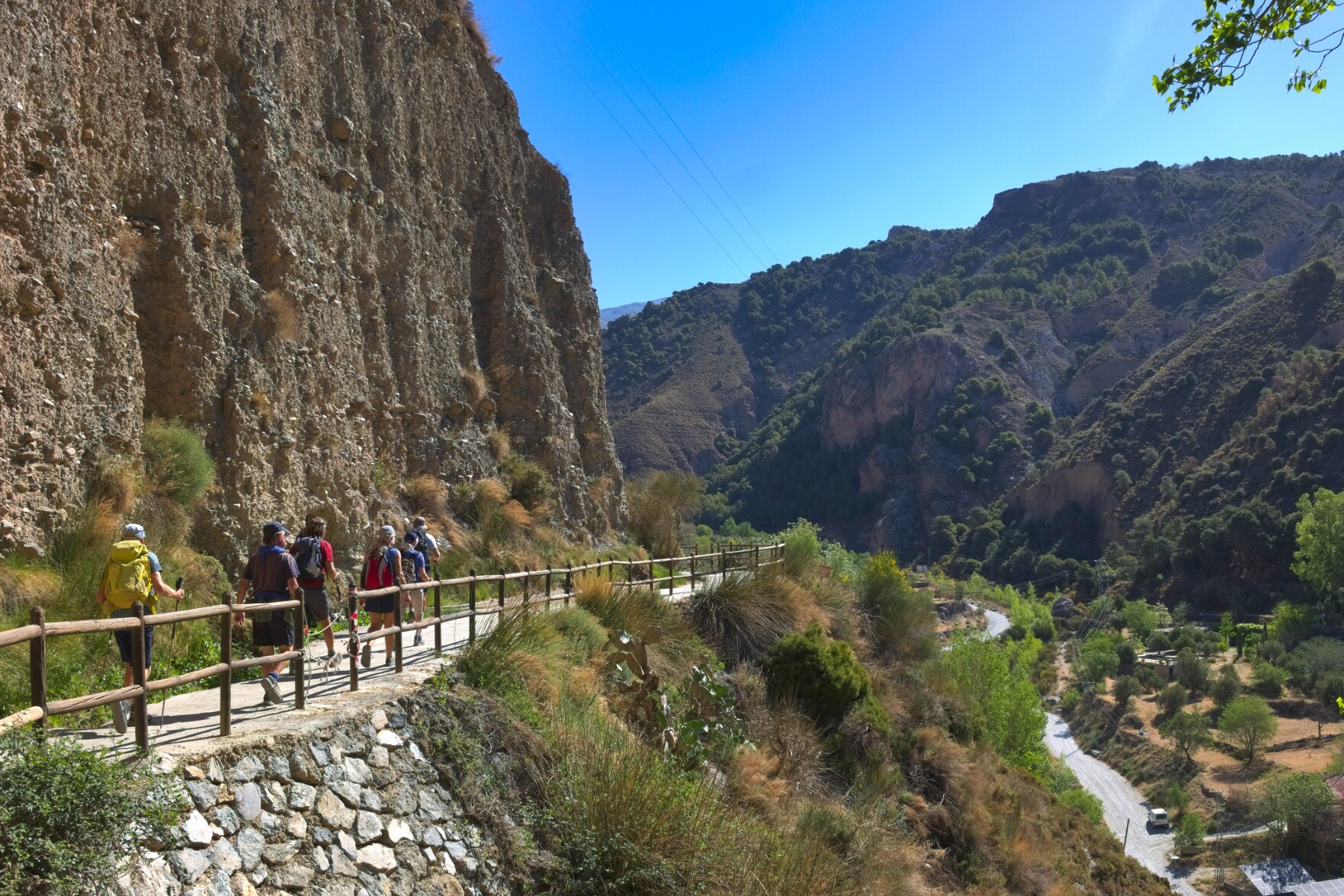 A group of hikers walk along a narrow path with a fence guarding the drop. A cliff looms high above
