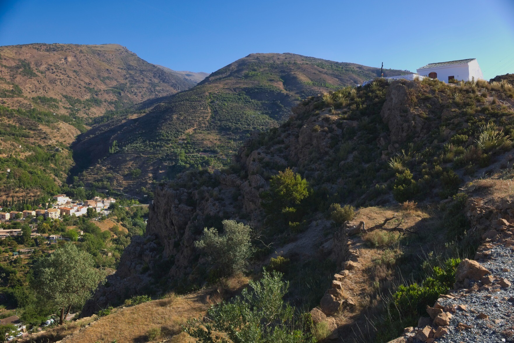 A rural spanish scene with the outskirts of a small town to the left. A deep valley runs up towards the mountains. On the right is a small chapel on a hill.
