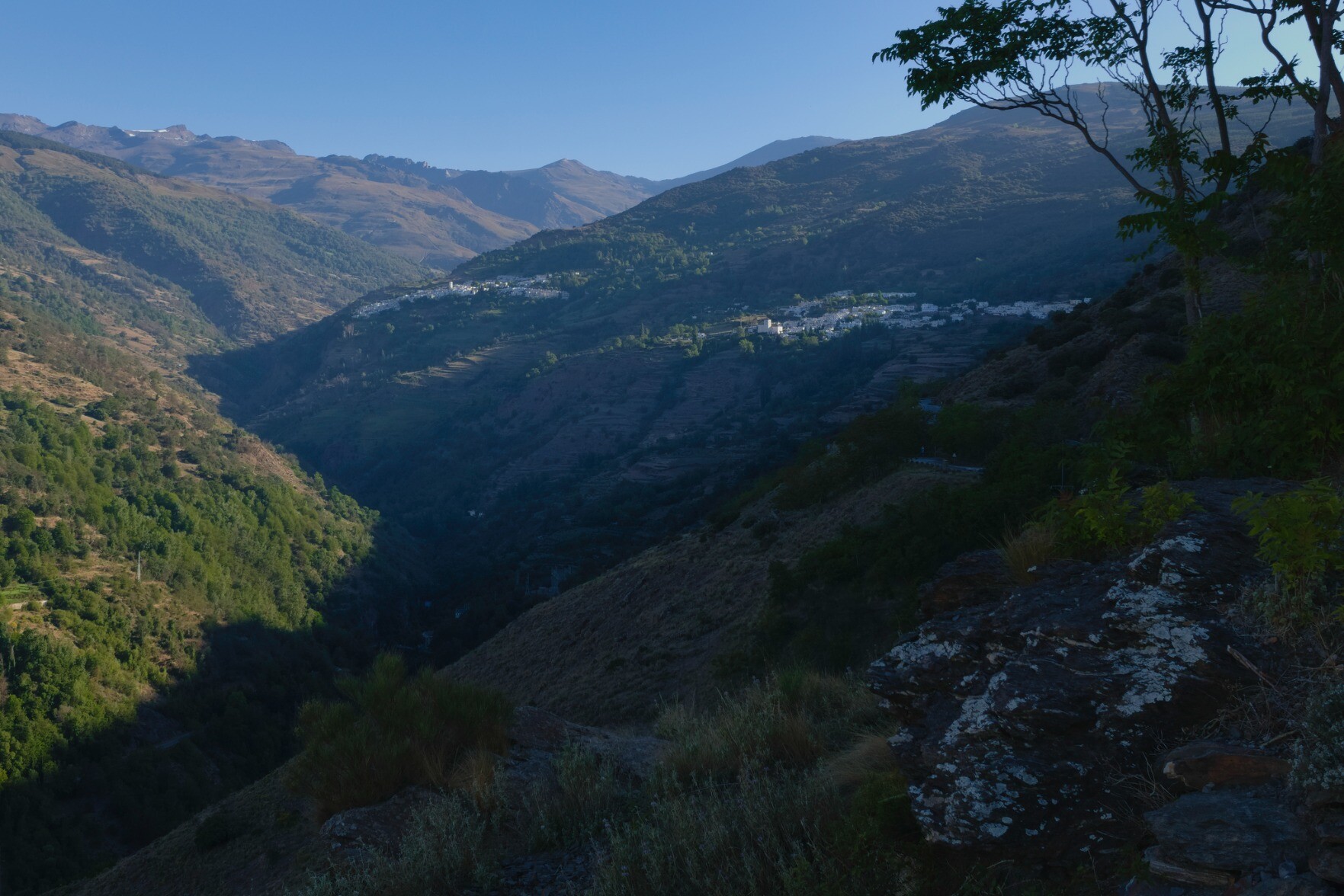 Two white hill villages appear in the morning sun. A deep valley to the left is in shade whilst further left the hillside is bathed in sunshine. In the distance some high mountains appear as a jagged line