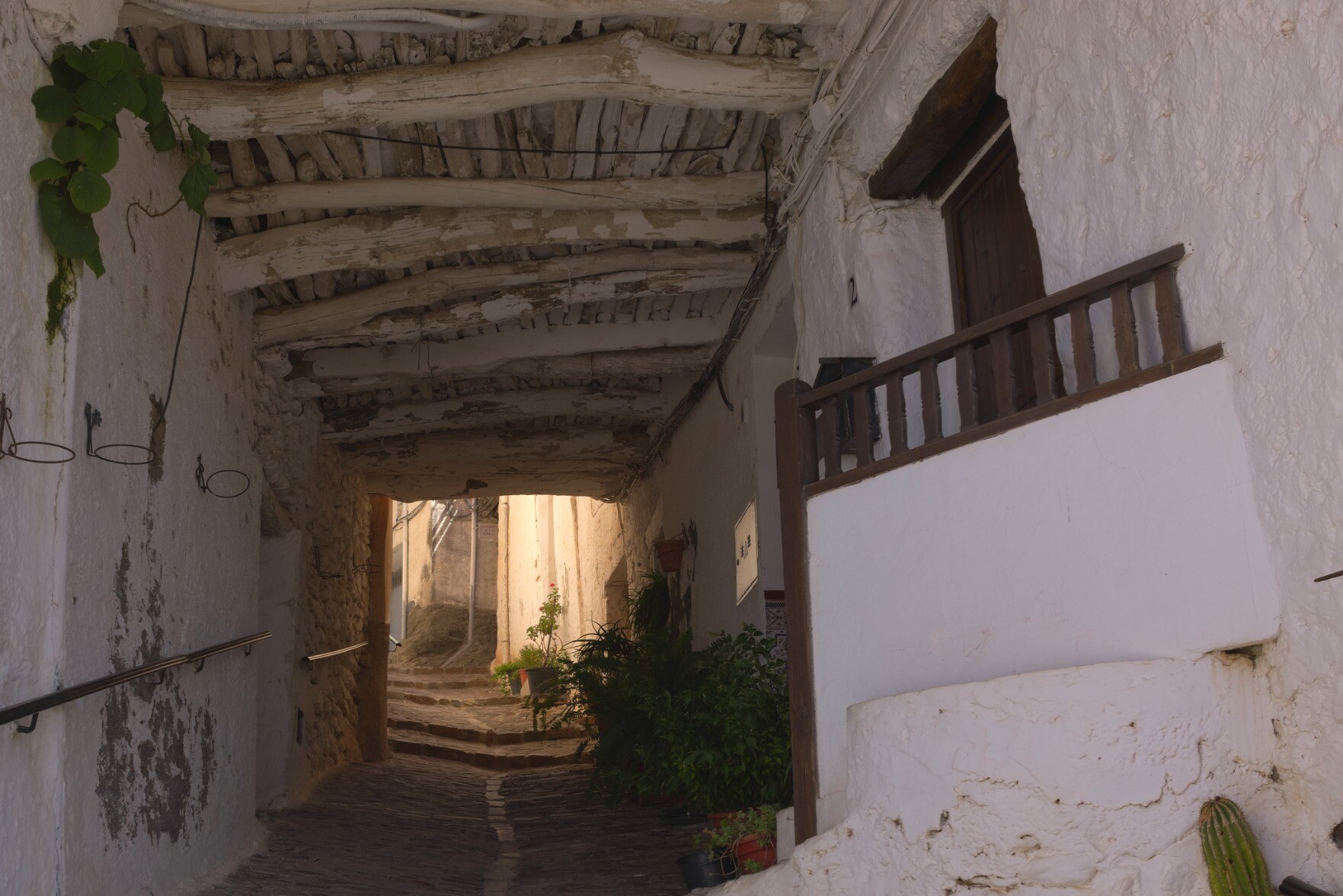 A narrow alleyway with old buildings on both sides leads to a sunlit exit. The roof is built with traditional materials 