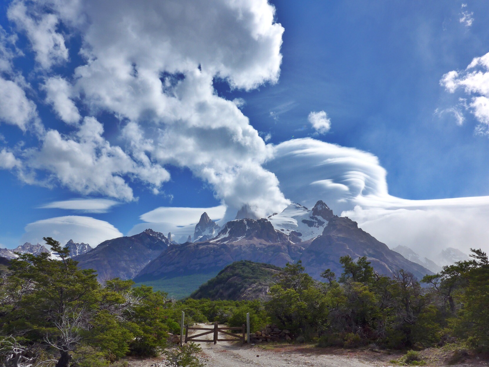 In the foreground a gate and dirt track lead to some green bushes. Behind rise some jagged mountains with huge circular lenticular clouds above them. Blue sky too