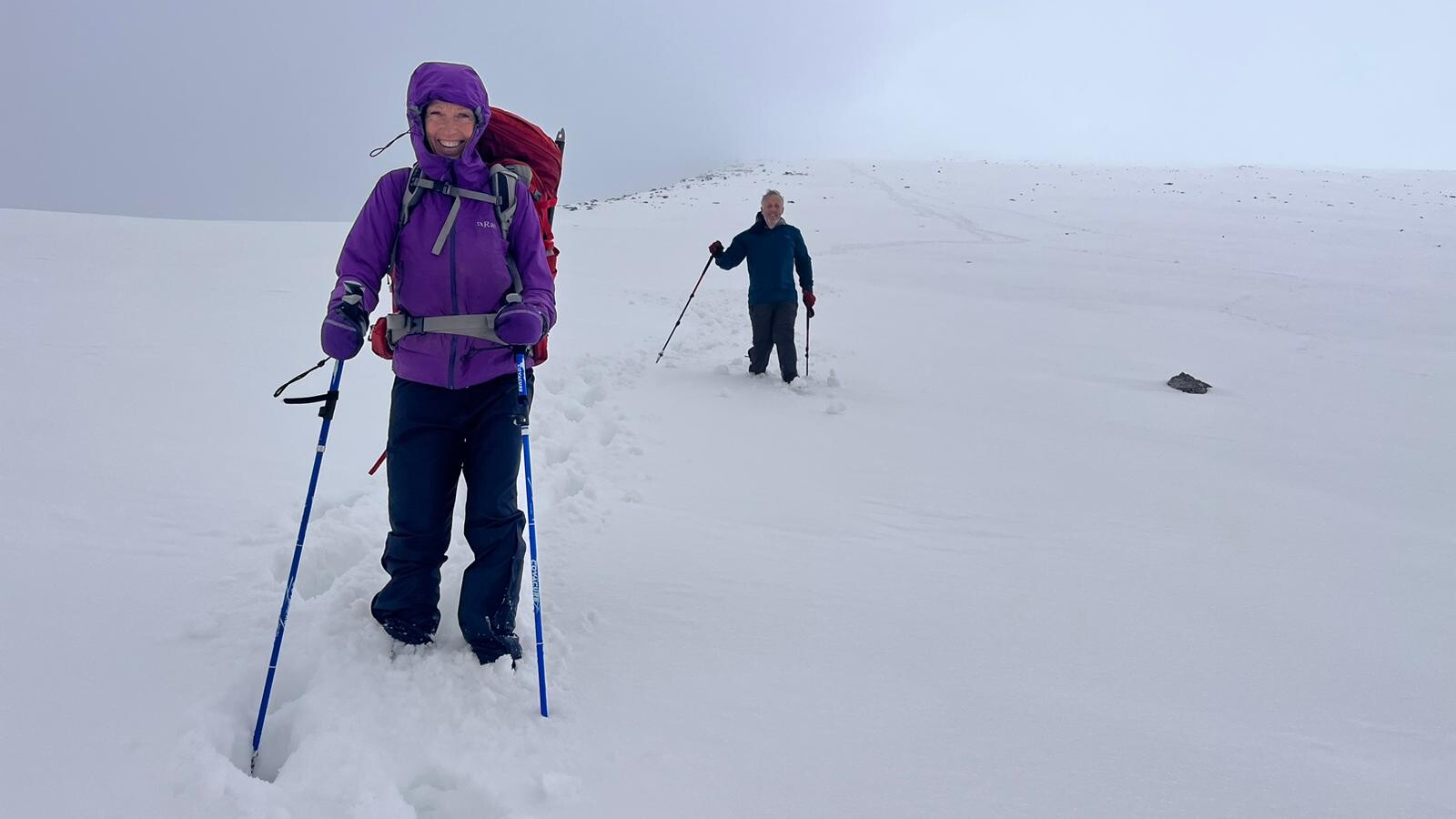 A lady in purple jacket is deep in snow. behind her a man dressed in black and tracks in the snow leading back uphill