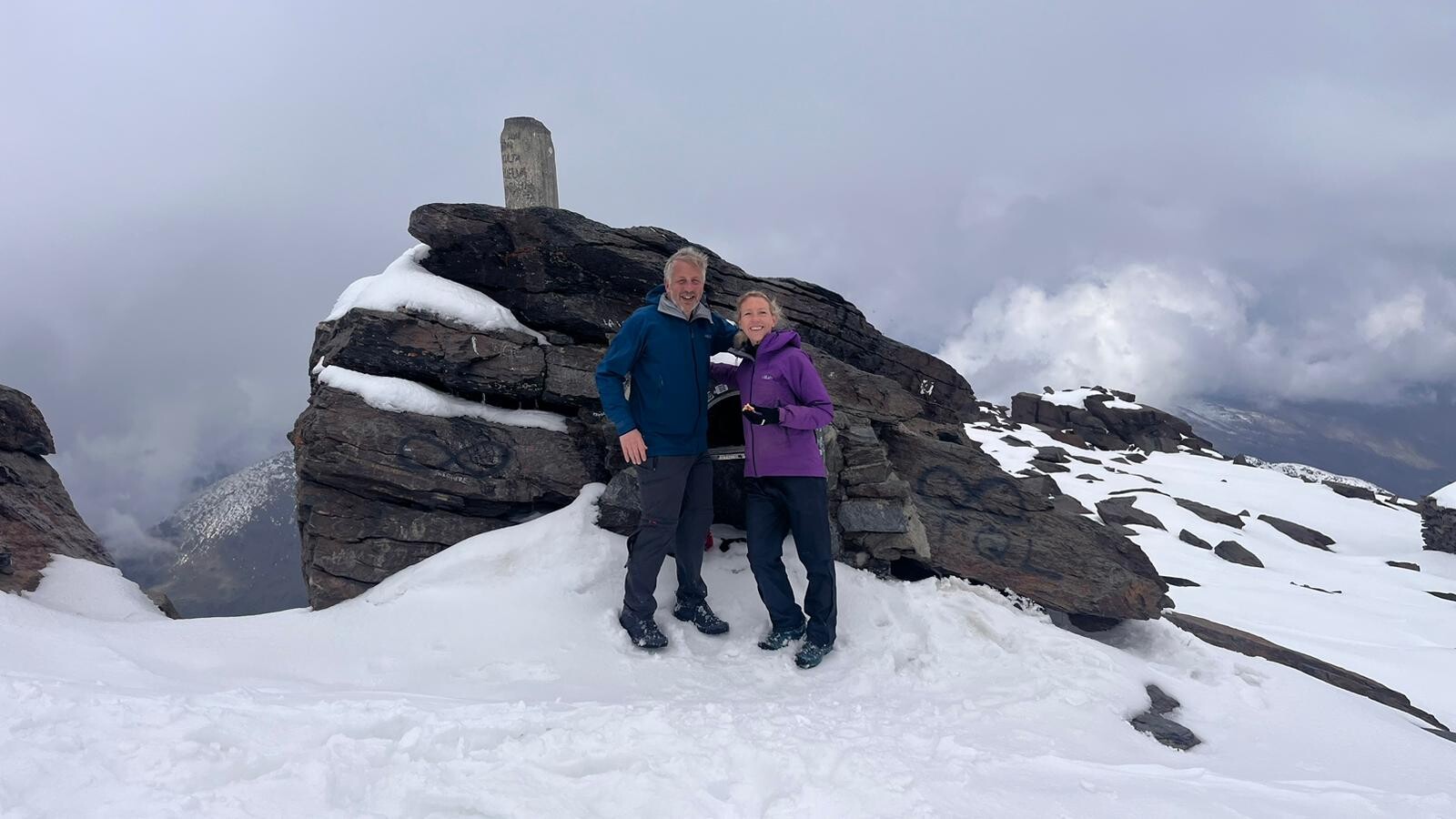 A couple of hikers on the summit of Mulhacen mountains. There is a large cairn. Its looks cold, there is snow on the ground and threatening clouds around