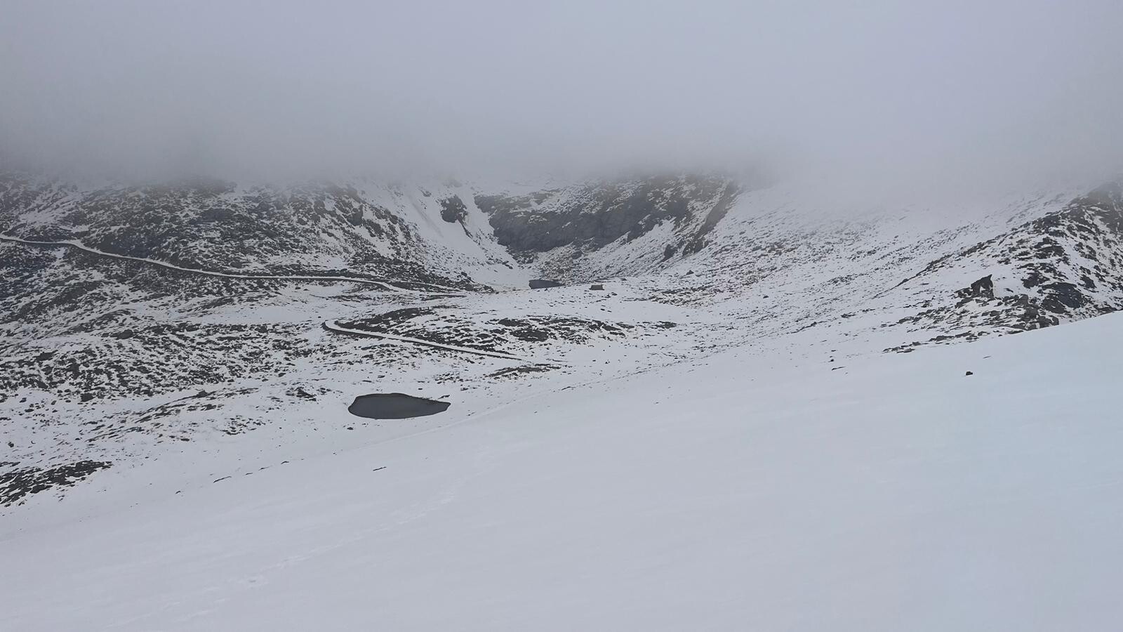 The small hut of the Refugio de Caldera can be seen nestled in a hollow surrounded by cliffs. There is snow all around and grey clouds above