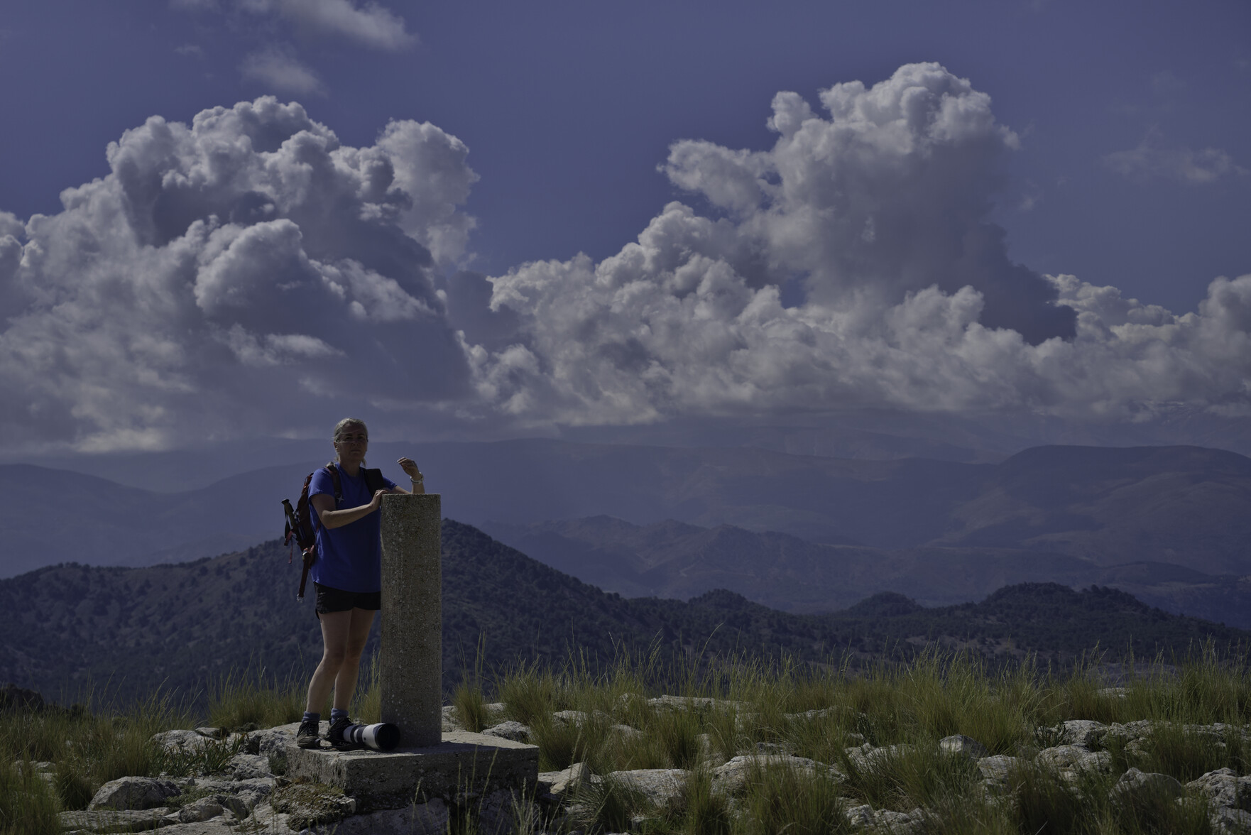A person stands at the summit of a mountain. Distant views to big peaks with large clouds building above them