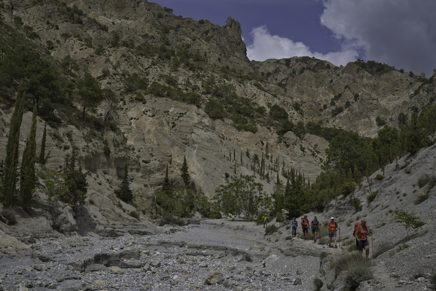 A group of walkers are coming down the right hand side of a wide but dry river bed. behind are steep cliffs that reach up into the mountains and have some trees at mid height