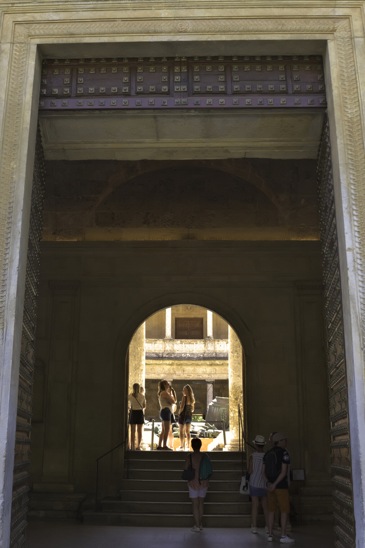 An imposing entrance gate leads into the Palacio de Carlos Quinto of the Alhambra Palace in Granada. A small group of people stand out in the sunlit interior.