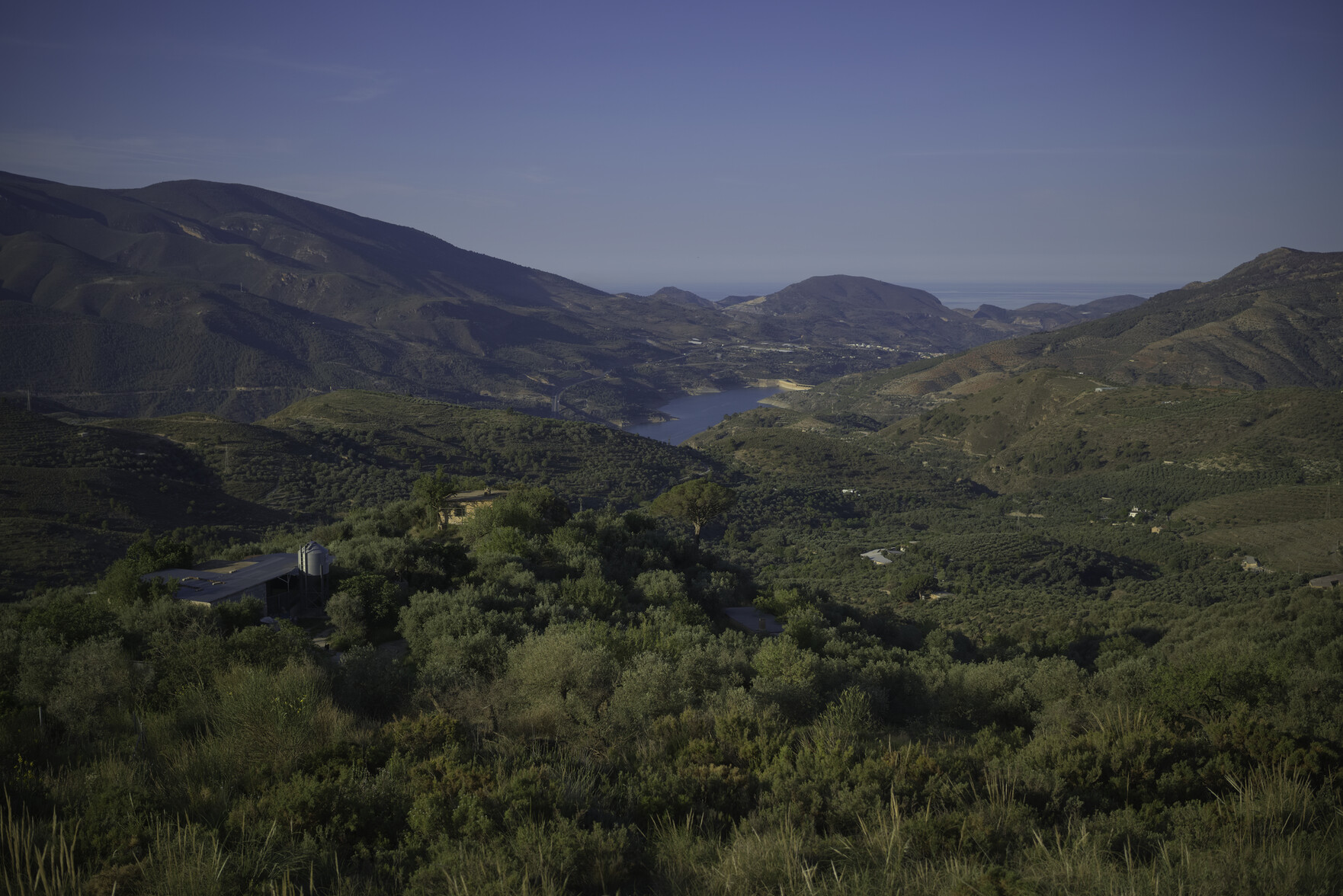 A view over green fields and trees towards a blue reservoir. To the left are high mountains and to the right the blue of sea and sky