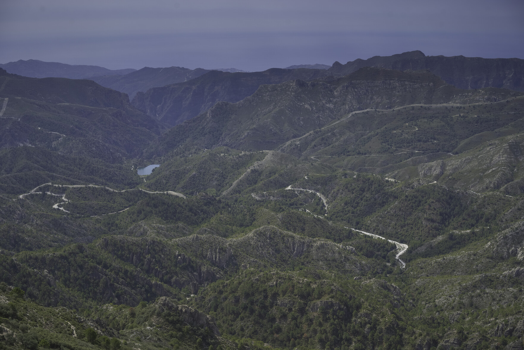 A view from the summit southwards. There is a complicated landscape of green forests and deep valleys. Some dirt tracks cross this terrain. In the distance is the Mediterranean Sea.