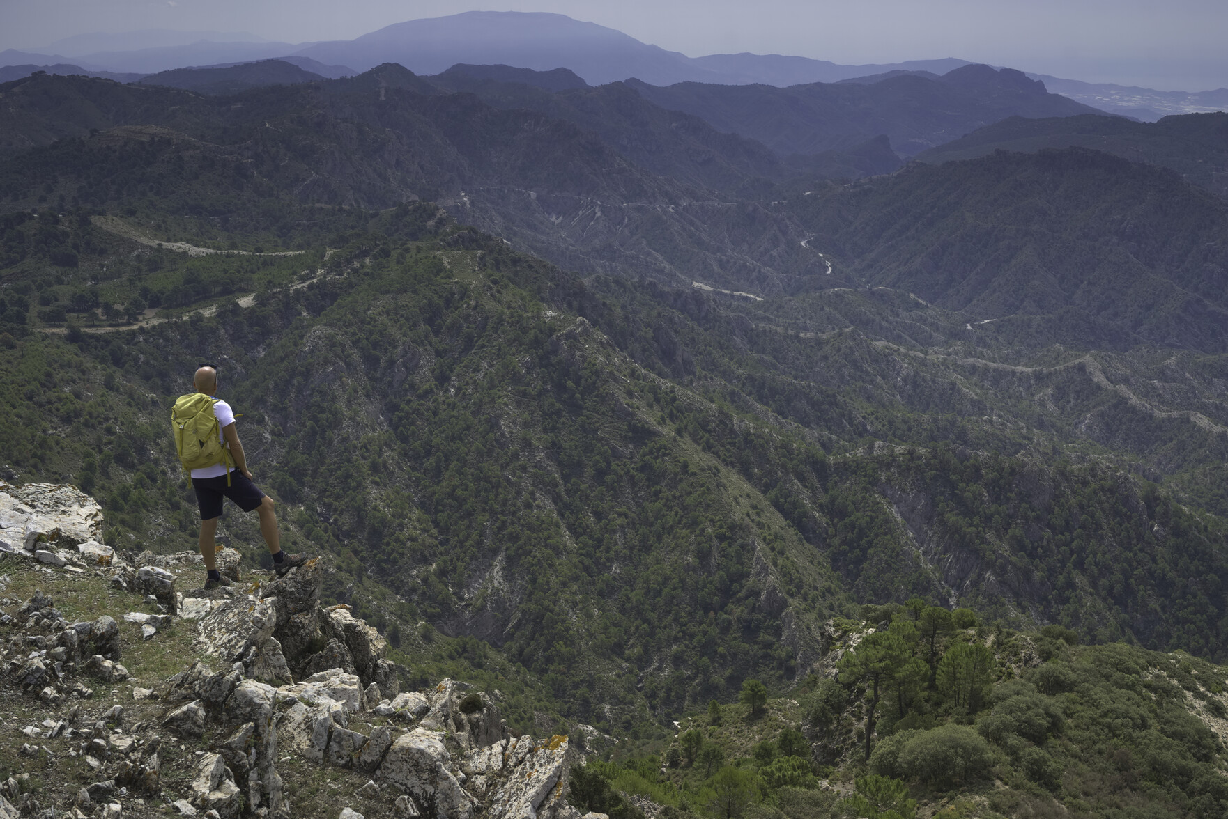 A man stands on the left of the photo. To his right is a complex mass of rough country, forests and dirt tracks that lead to distant mountains