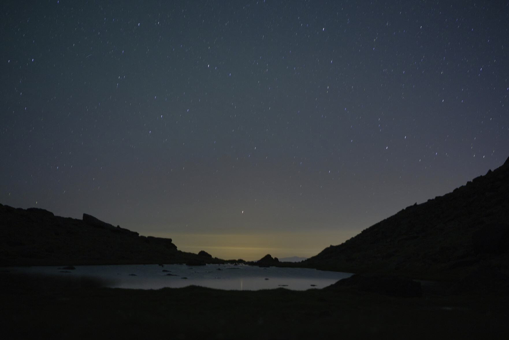 The night sky is full of stars. There is an orange glow lower down and the reflections in a mountain lake