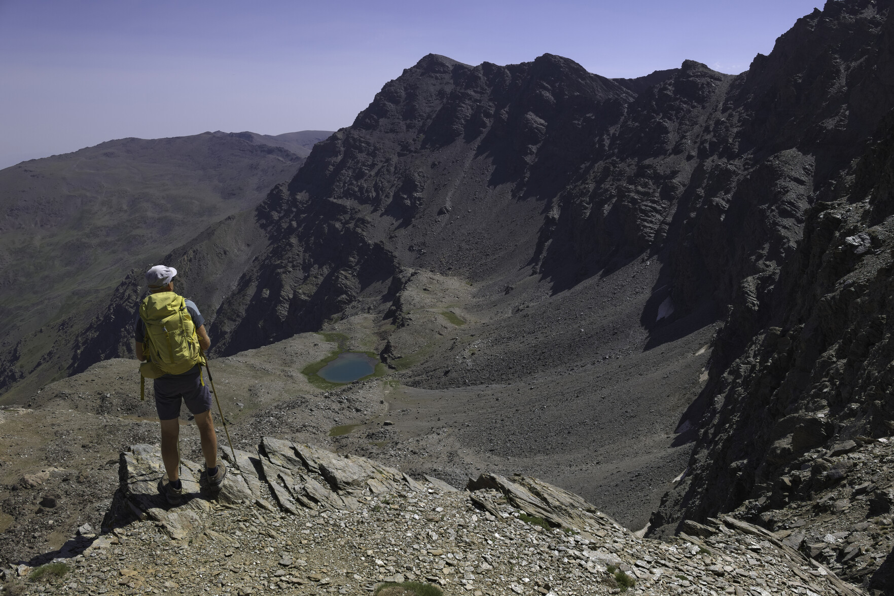 A person with a yellow rucksack stands looking over a drop towards a distant lake. To the right almost vertical cliffs