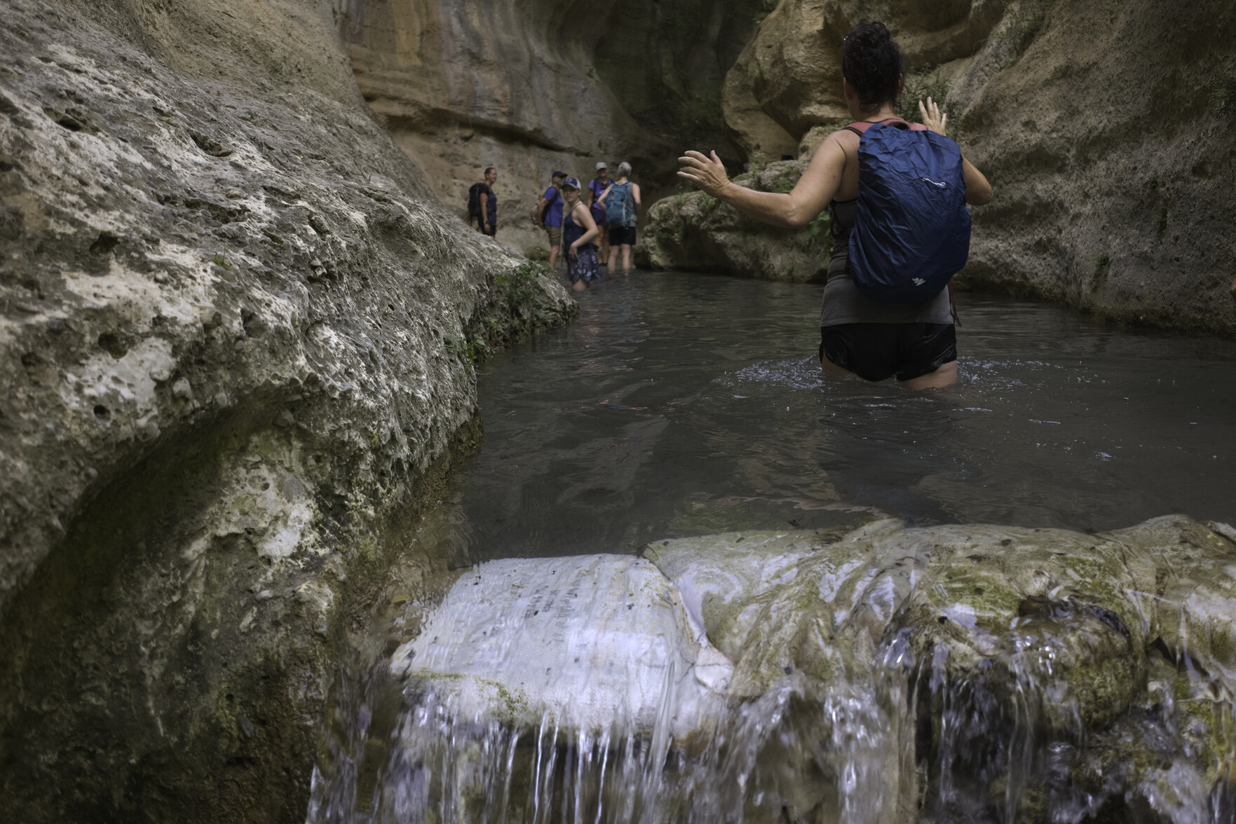 People wade through waist deep water in a small lake formed by a steep sided, dramatic gorge