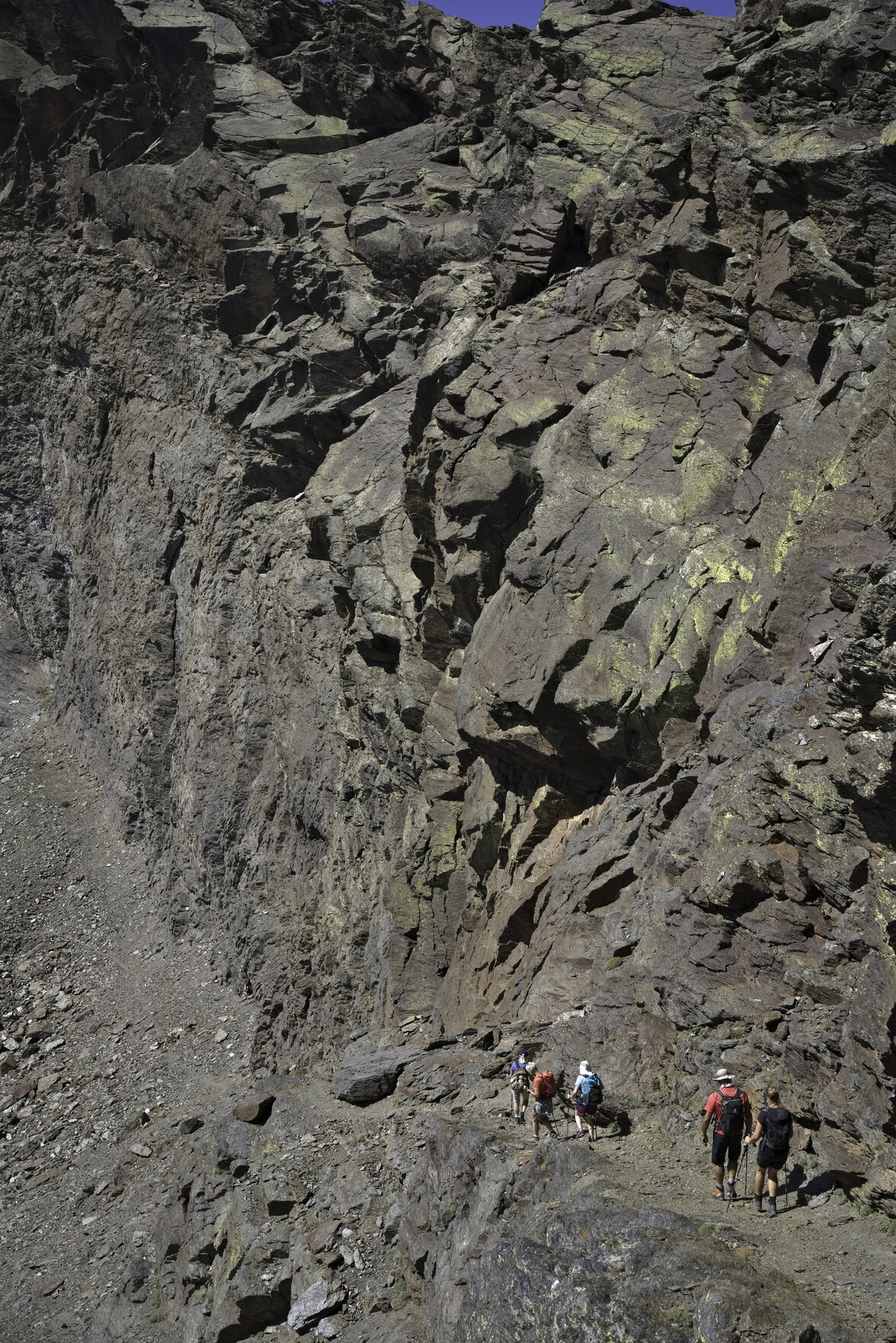Huge rock walls tower above some diminutive hikers walking down a path at the bottom of the photo