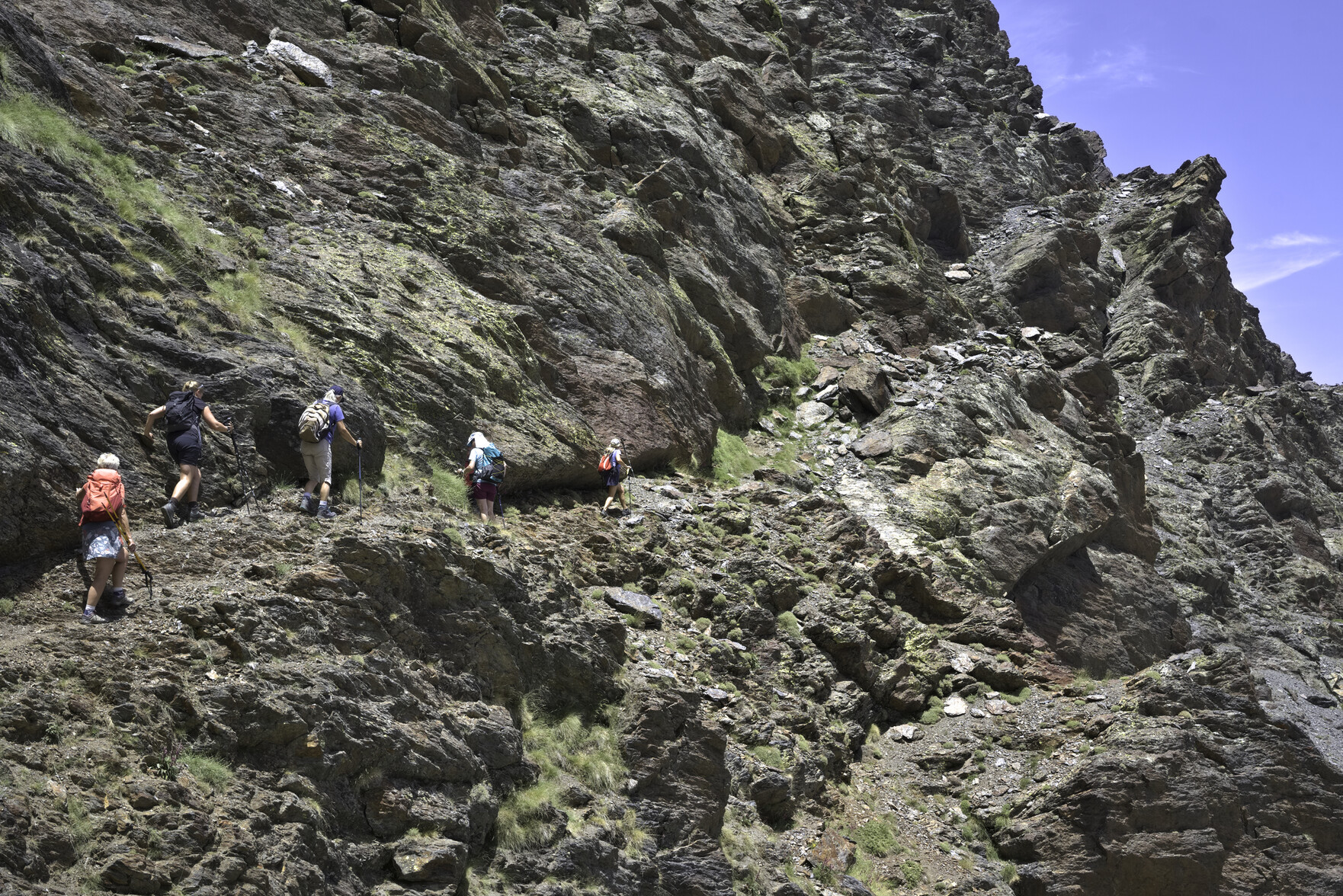 A group of hikers on the left pass along a narrow path that traverses up across the face of a steep mountainside covered with cliffs