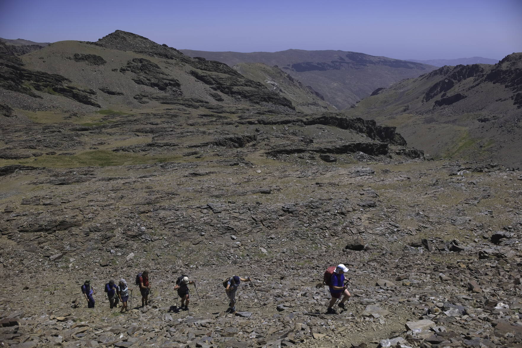 Hikers toil in the hot sun up a barren semi desert like landscape. Blue sky above