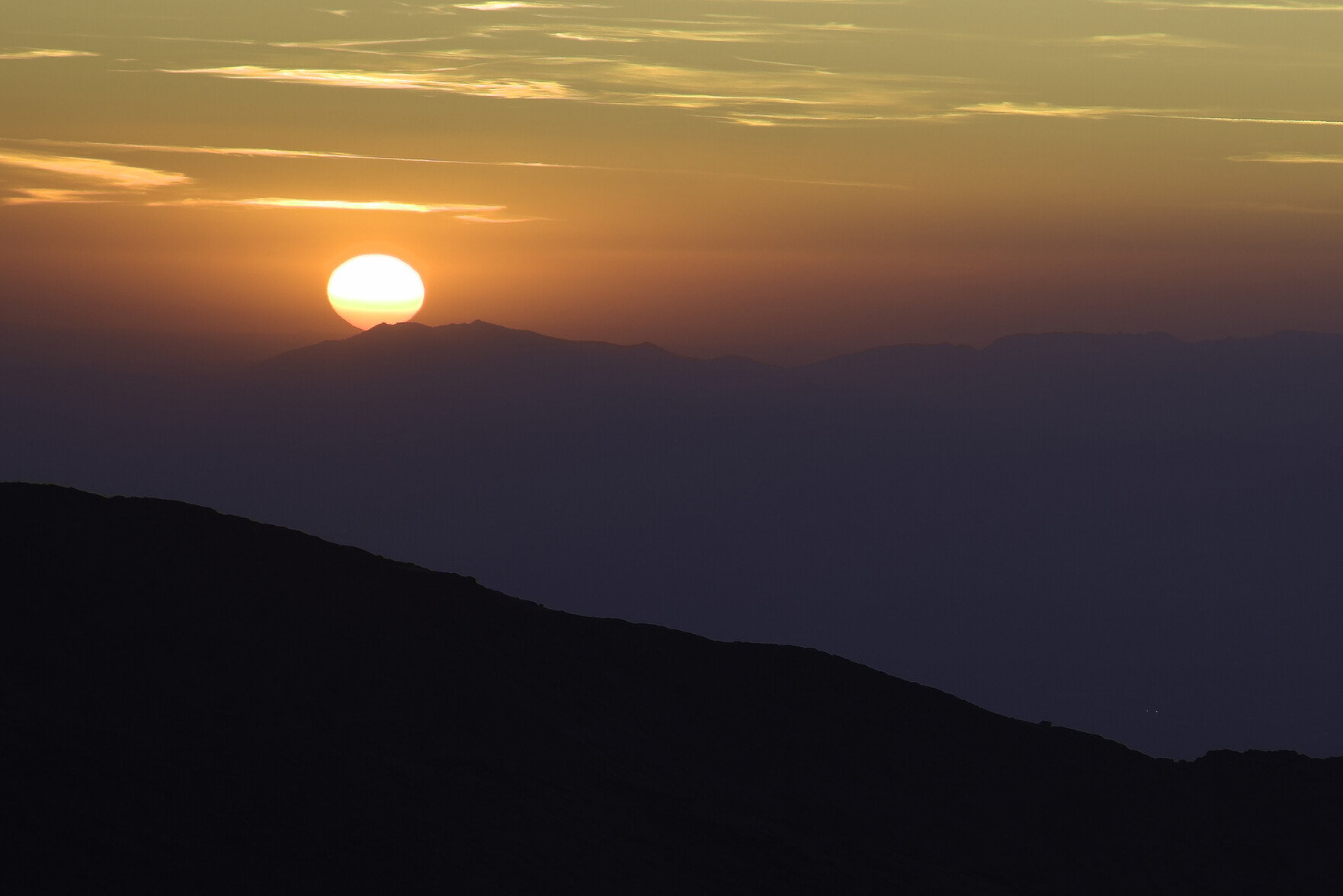 The sun is setting over some hills. Very dark or black foreground but various orange shades around the sun