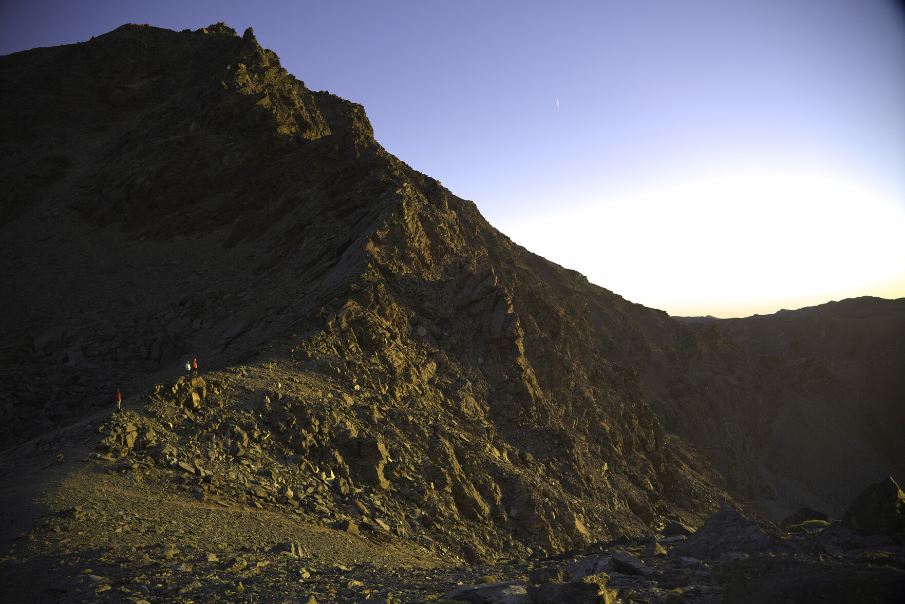 A group of people illuminated in the evening sunshine watch the sunset. behind rises a spiky mountain ridge