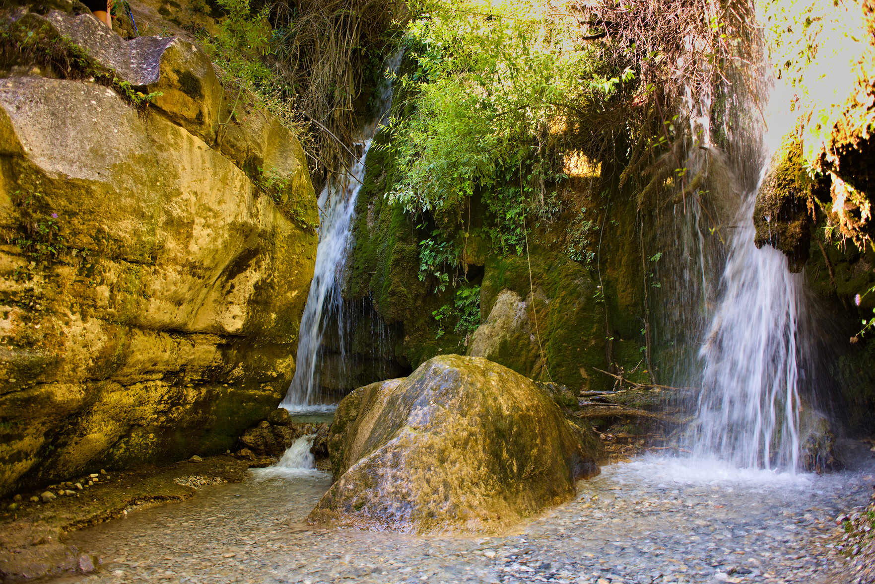 Two waterfalls plunge down from green vegetation  at the top of the image. A large rock is on the left. Below a small lake forms