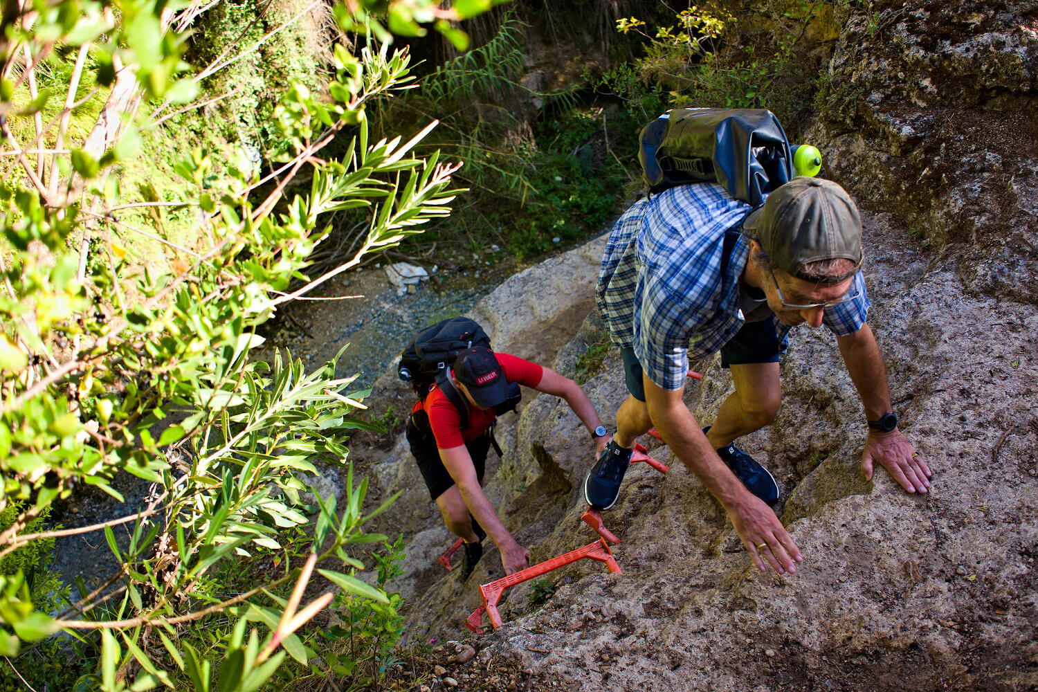 Two hikers climb some steps up a rock face below a river