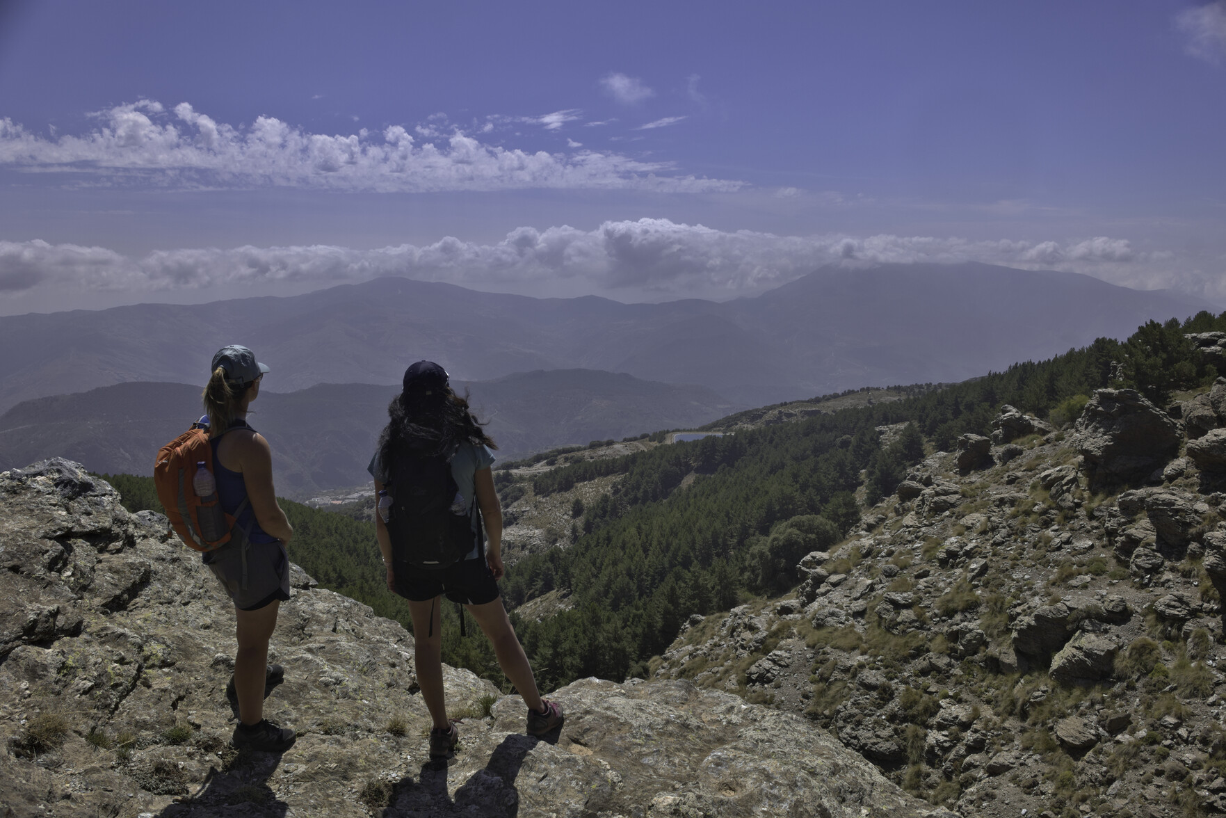 Two ladies stand on an outcrop of rock to the left. Clouds start to build over the mountains behind