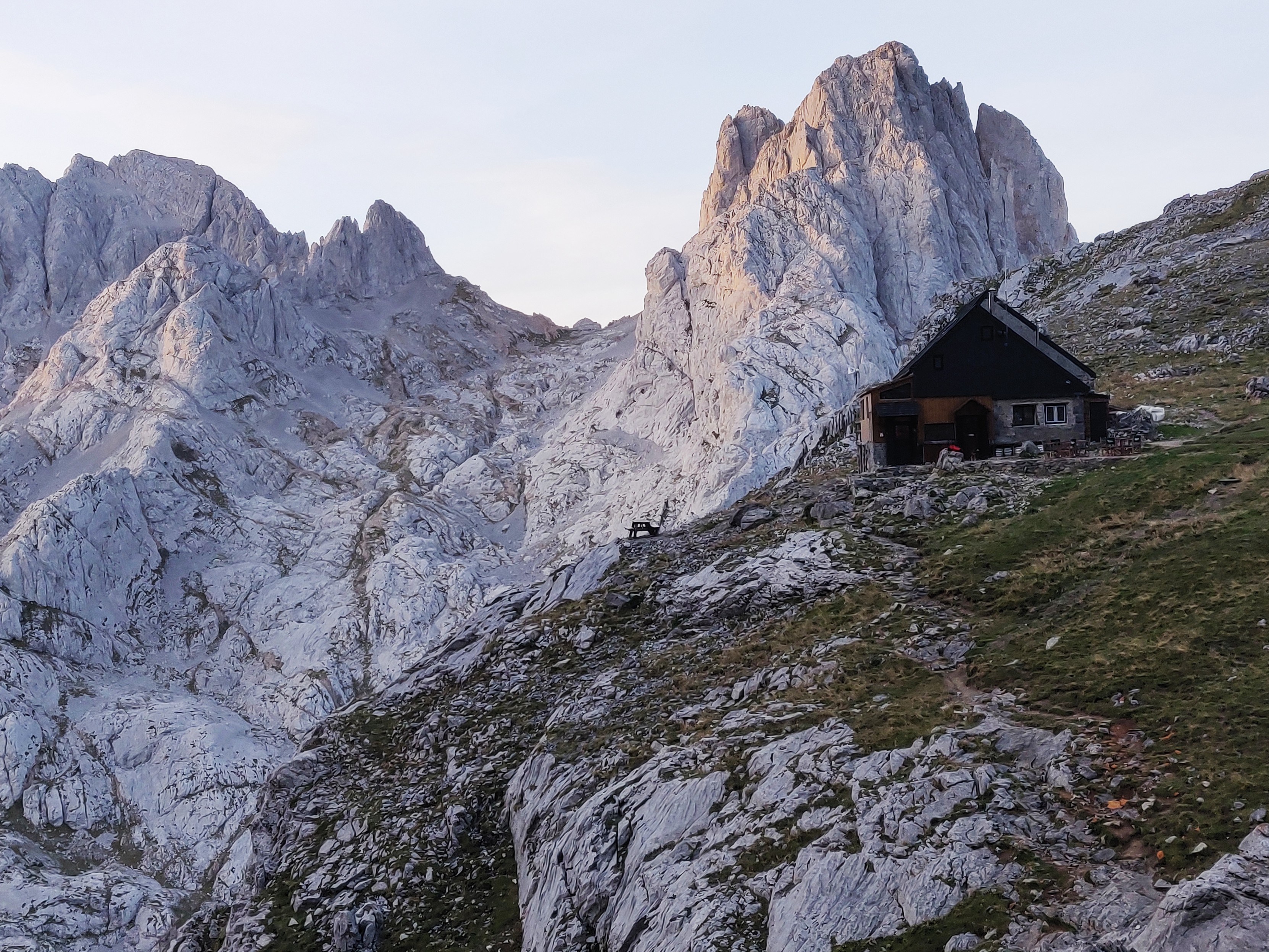 A small mountain refuge sits on a grassy shelf to the right. Behind lie some grey rocky peaks