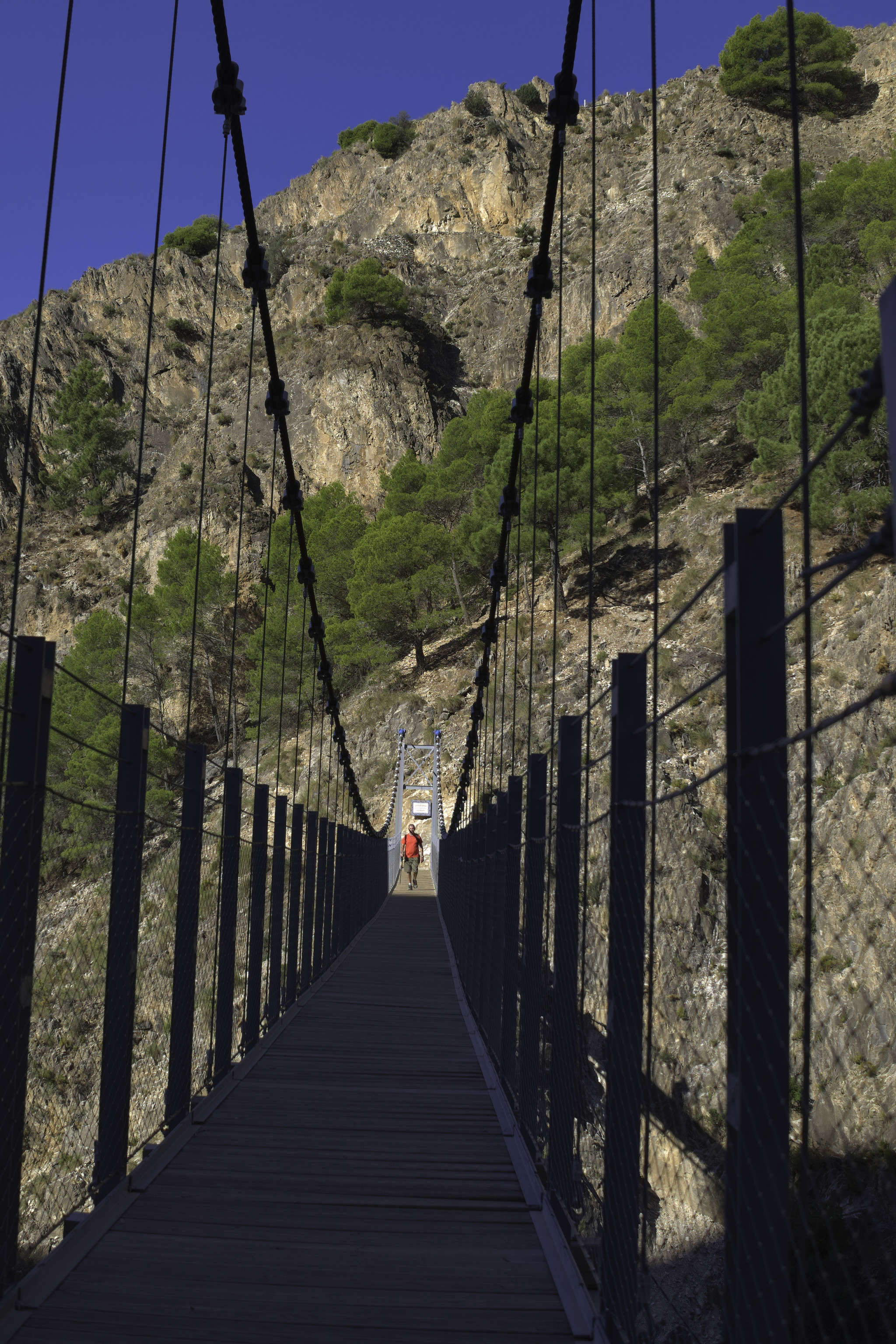 A huge hanging suspension bridge in shadow over a gorge with a hiker crossing in the sun