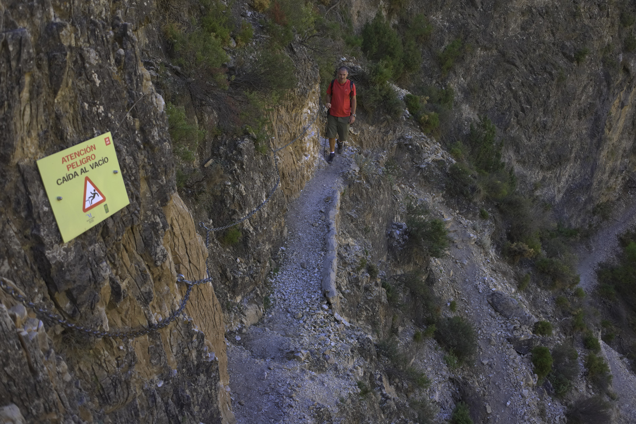 A man in red walks a narrow path with a chain to assist. At the end is a sign indicating danger of falling in spanish