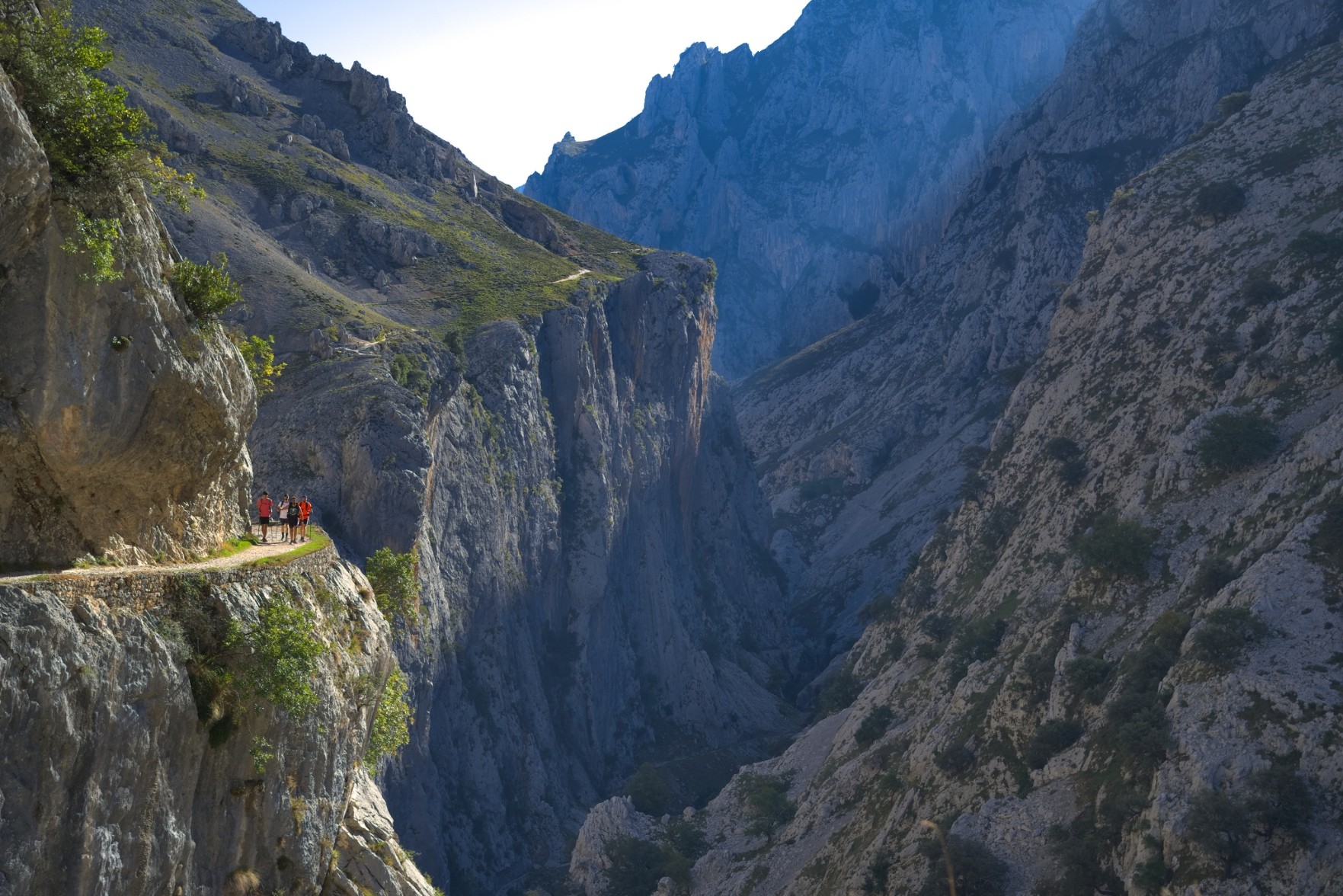 Hikers on a sunlit narrow path high on the left above a deep gorge in shadows