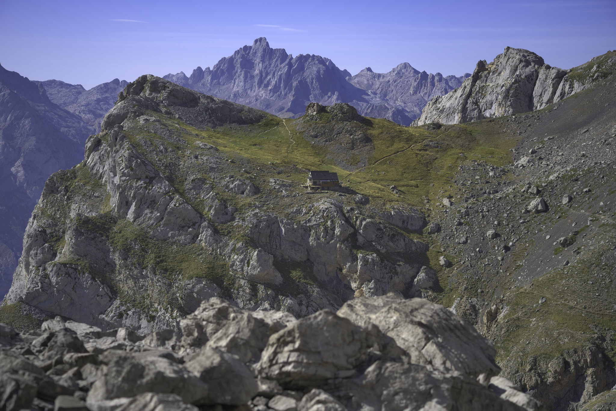 A small mountain refuge sits on a grassy shelf surrounded by steep cliffs and high mountains